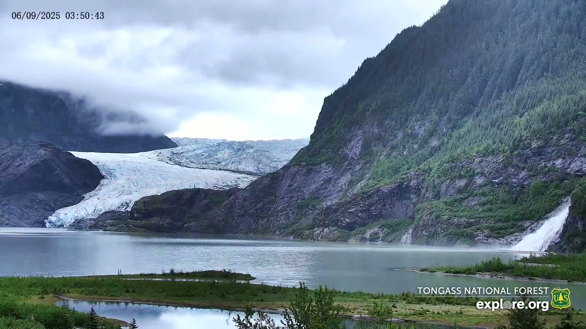 A glacier flows into a calm lake surrounded by steep, lush green mountains under a cloudy sky.