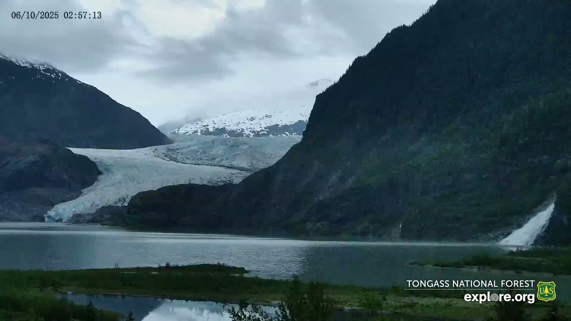 A glacier flows down a mountainside into a calm lake under an overcast sky, with a waterfall cascading down the mountain's cliff face.