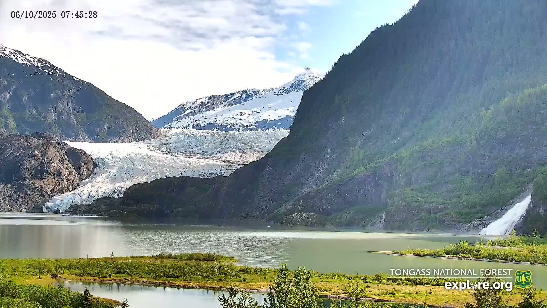 A glacier flows into a calm lake nestled between snow-capped mountains and a verdant forest, under a partly cloudy sky.