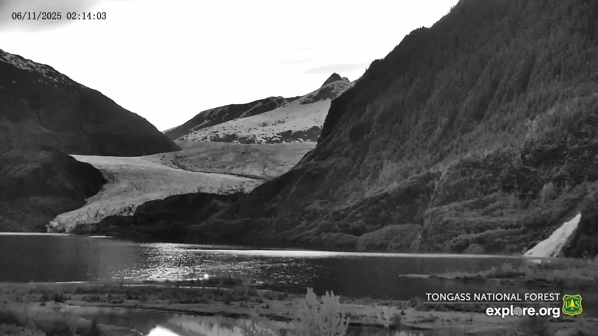 A glacier is visible between dark mountains next to a still body of water under a cloudy sky.