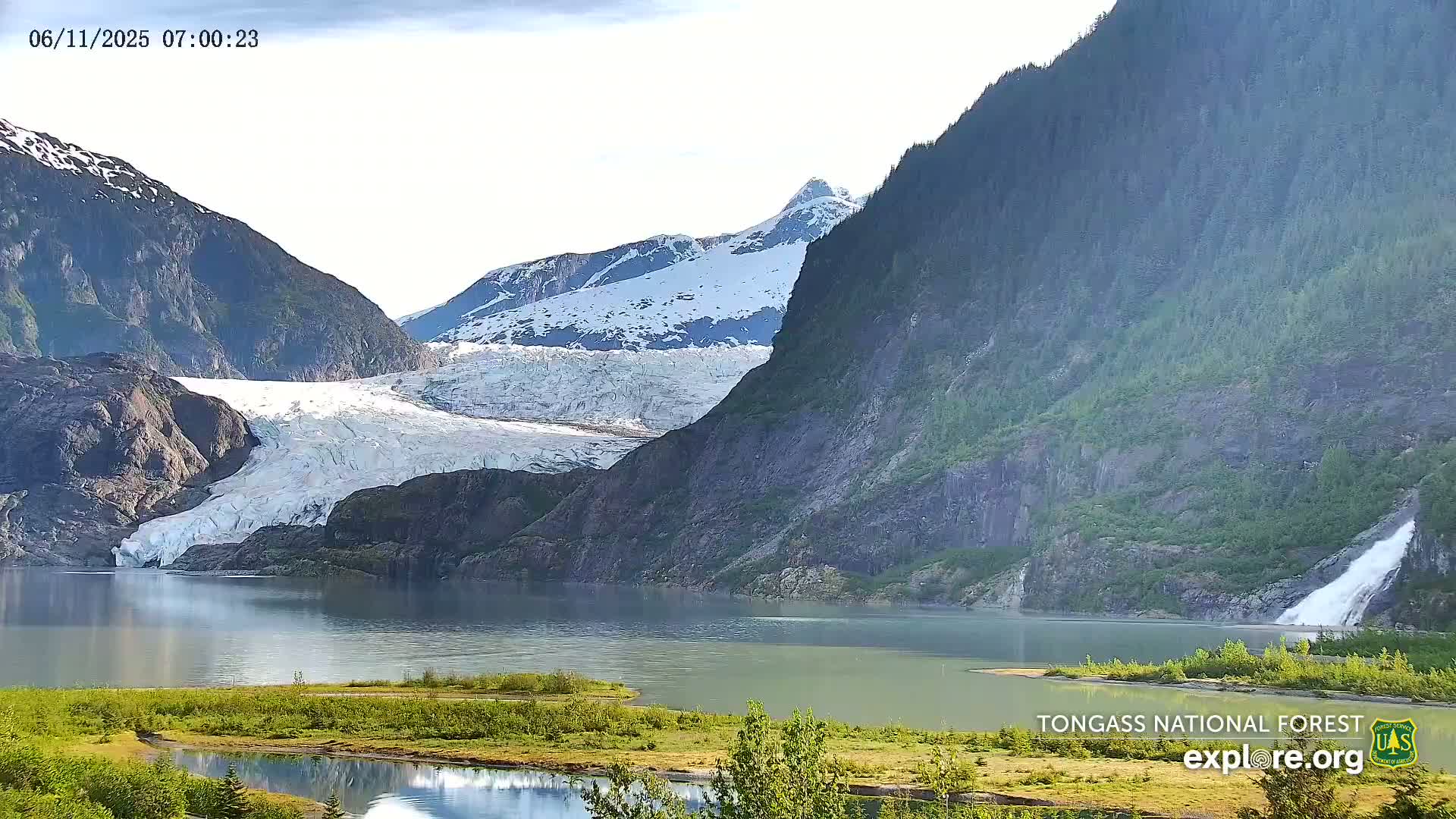 A large glacier feeds into a calm lake nestled between steep, snow-dusted mountains under a mostly clear sky.