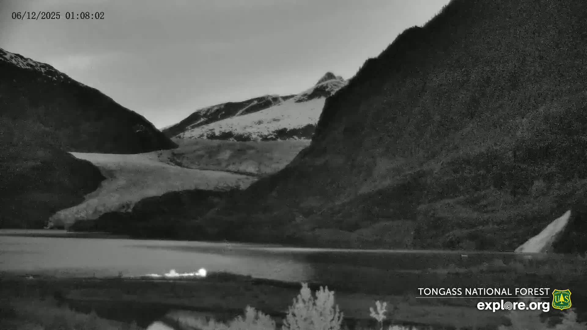 A snow-covered mountain range is reflected in a dark, still body of water under a gray, pre-dawn sky.