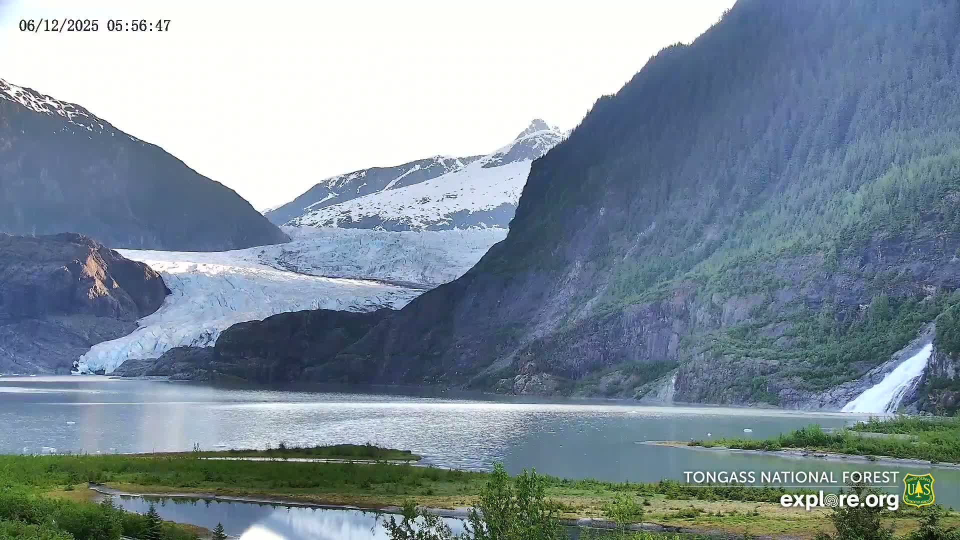 A glacier flows into a calm lake nestled between steep, snow-capped mountains under a bright, mostly clear sky; a waterfall cascades down a mountainside into the lake.