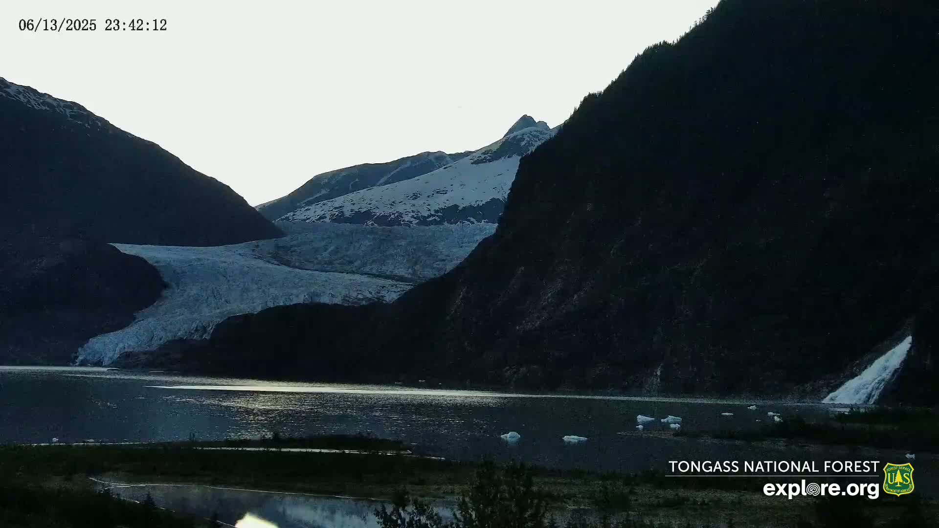 A glacier flows down a mountain into a dark, still lake under a twilight sky.