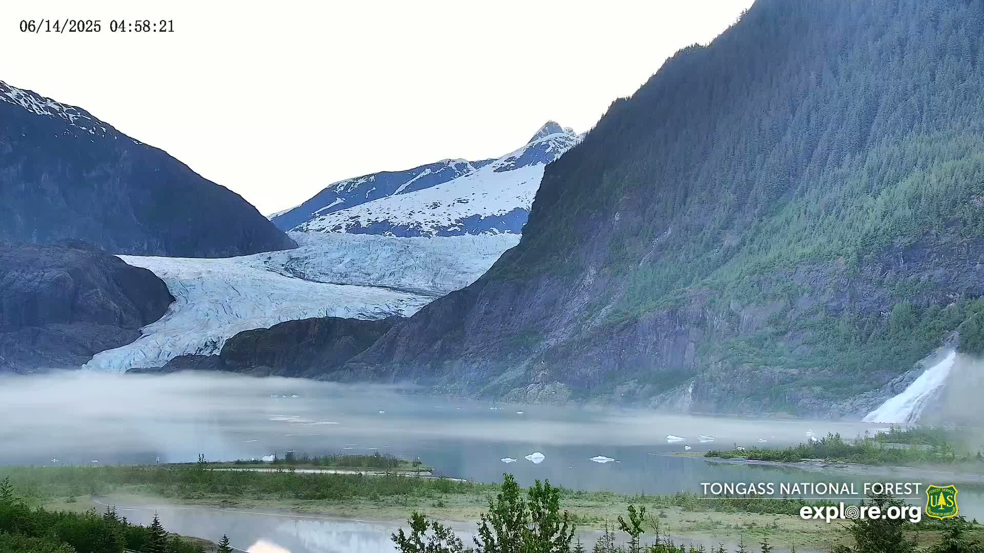 A large glacier is partially obscured by fog that sits on a calm body of water at the base of steep, snow-capped mountains.