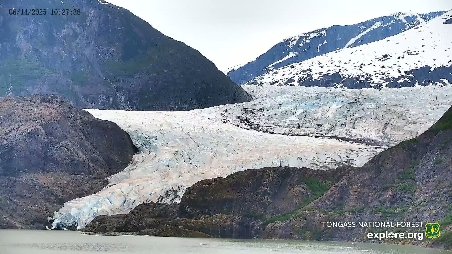 A large glacier flows down a mountain valley between dark, rocky hills on an overcast day.