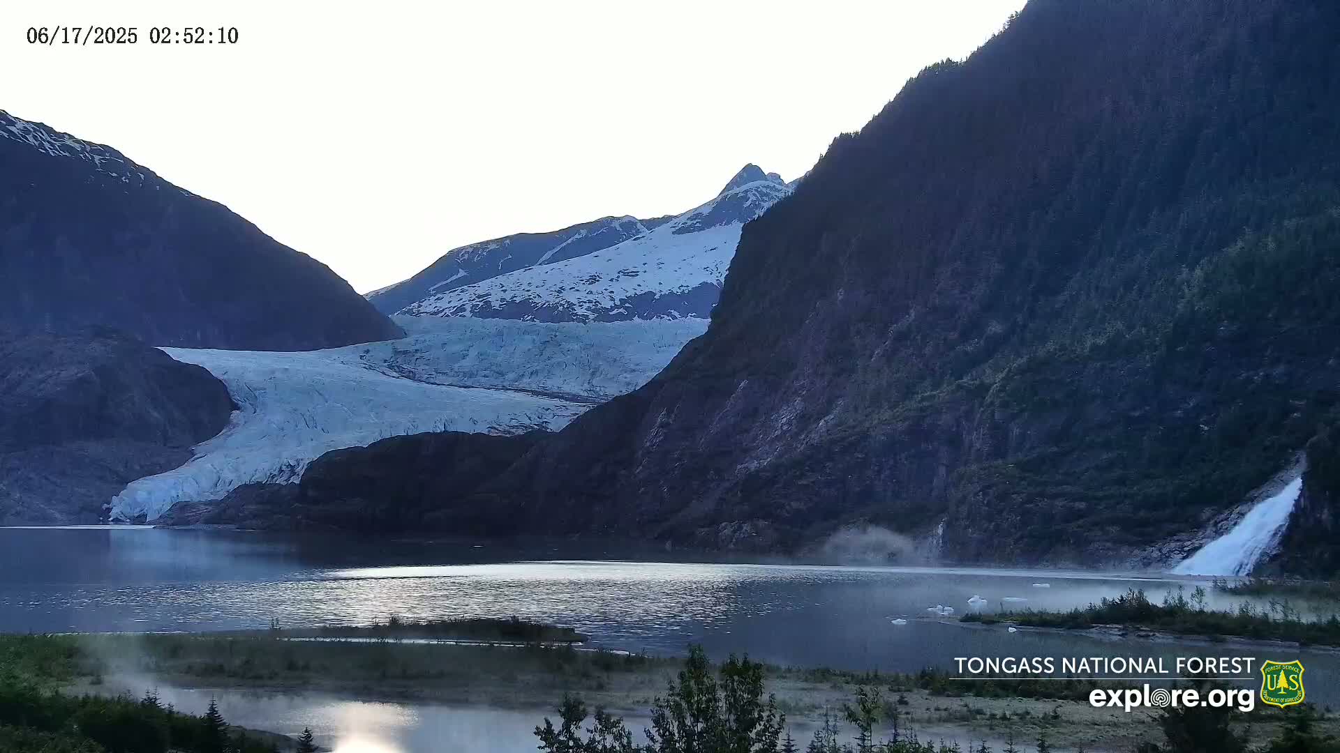 A large glacier flows down a mountainside into a calm lake, with a waterfall cascading down a cliff face on the right, under a bright, clear sky.
