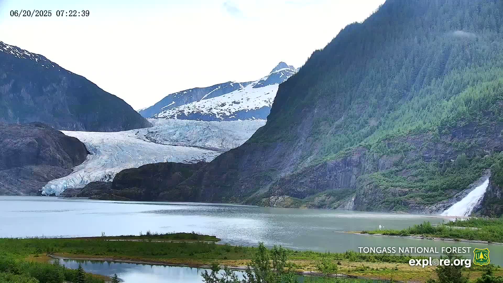 A glacier flows into a calm lake nestled between steep, snow-capped mountains on an overcast day.