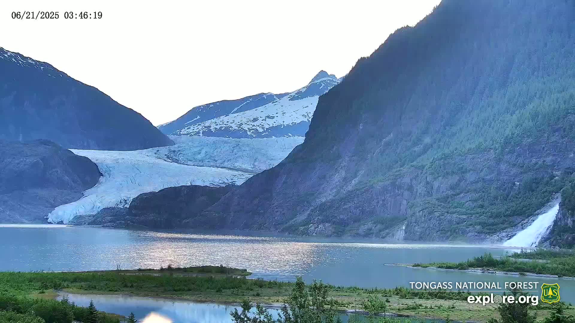 A large glacier flows into a calm lake, nestled between steep, snow-dusted mountains under a bright, clear sky.