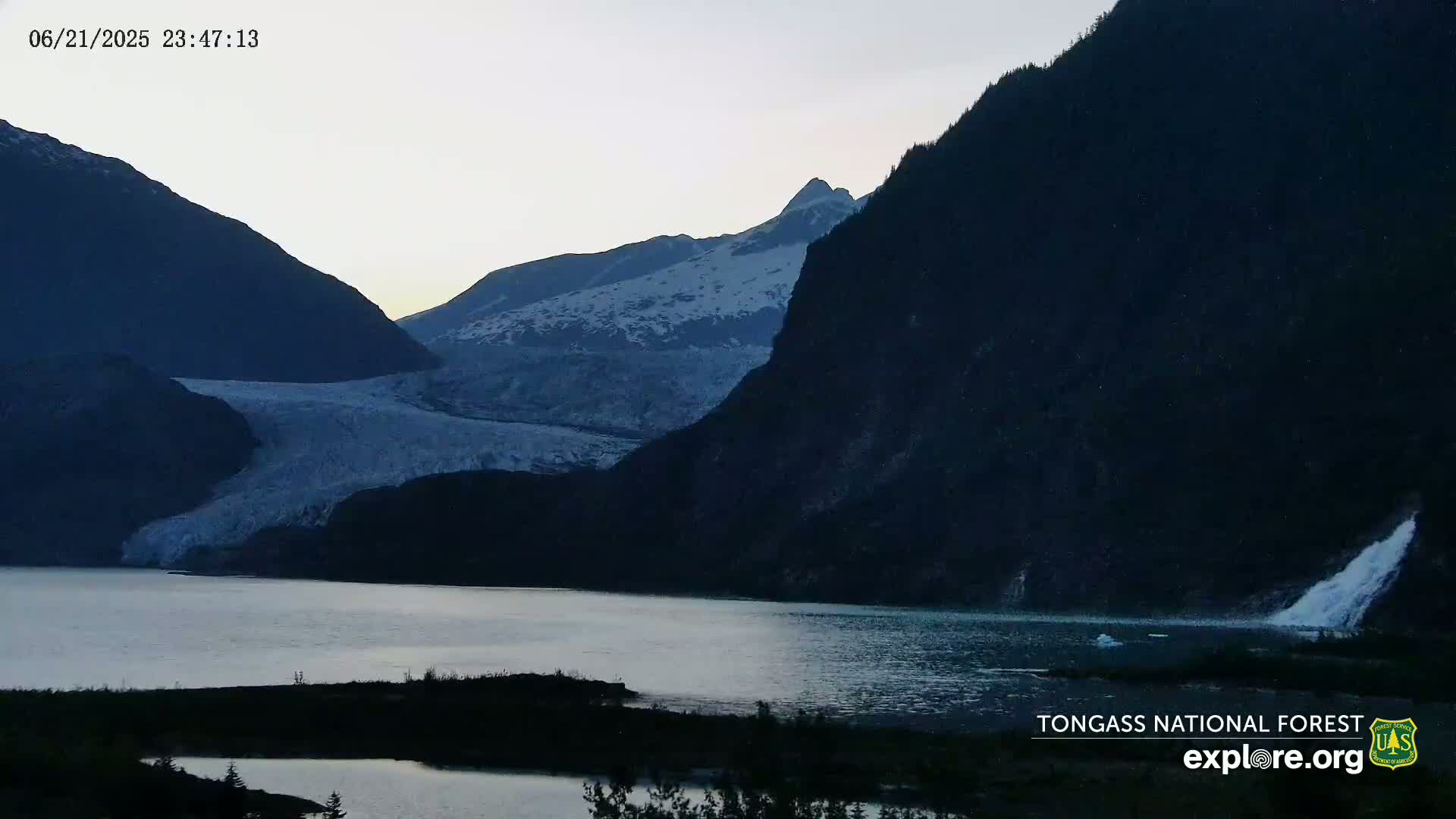 A glacier flows down a mountain into a calm lake at dusk, with a waterfall cascading from a cliff on the right.