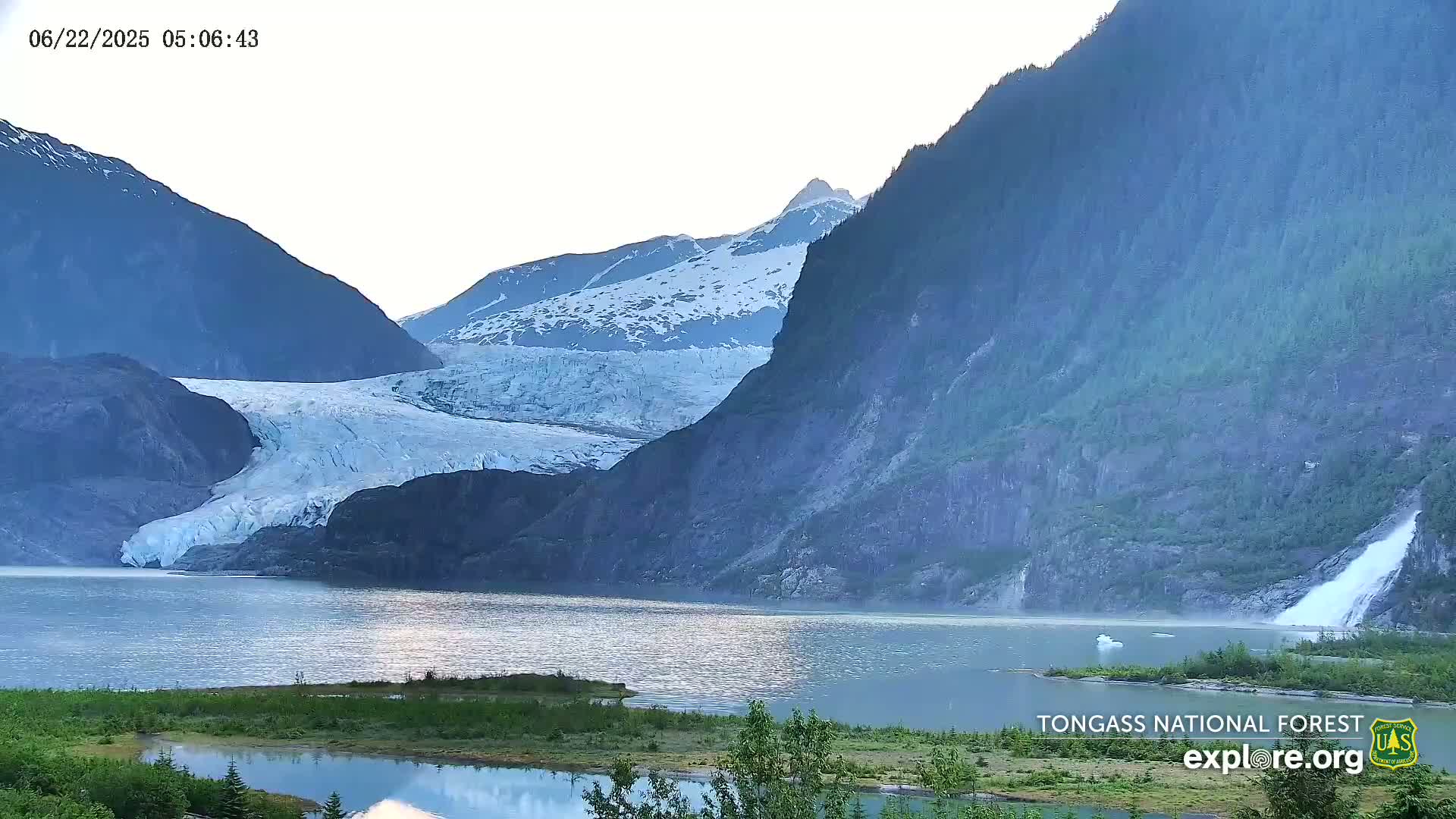 A large glacier flows into a calm lake nestled between snow-capped mountains on a partly cloudy day.