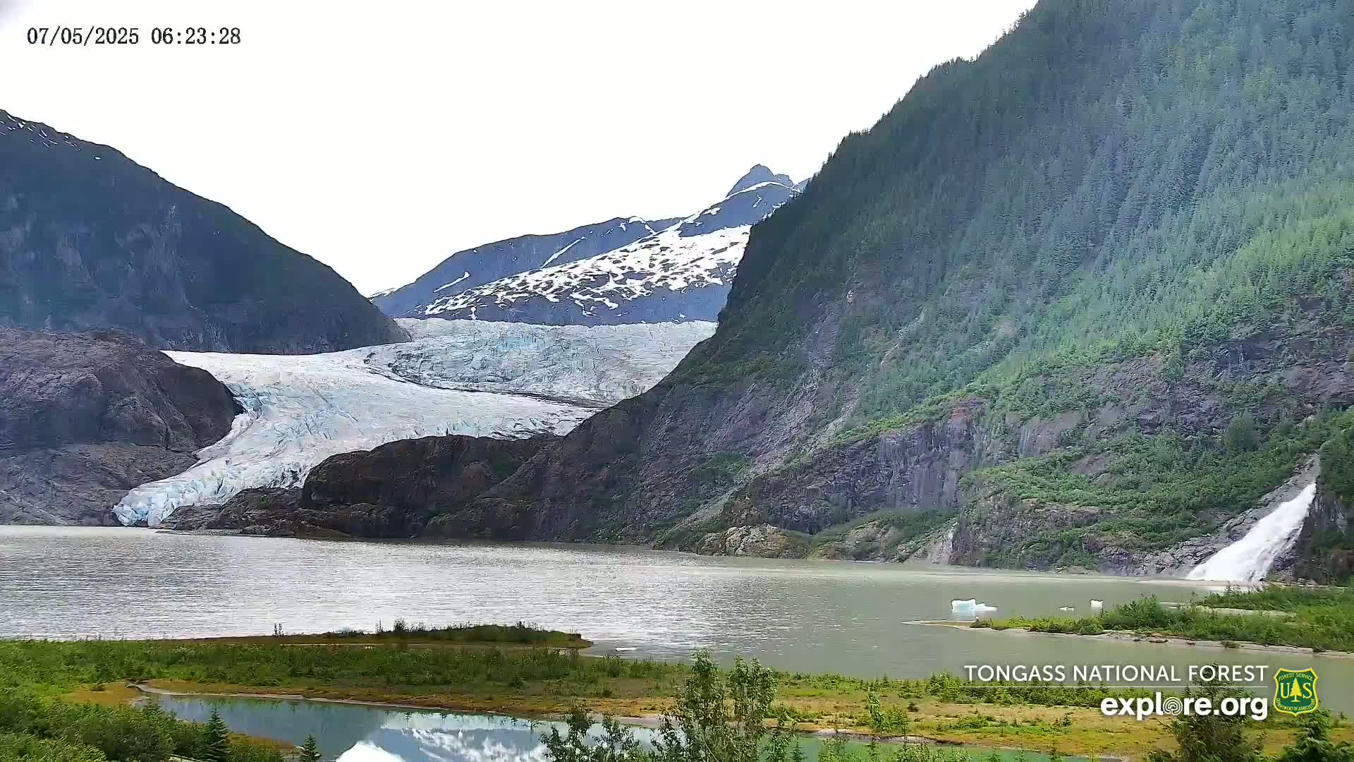 A glacier flows into a lake at the base of a snow-capped mountain and a lush green forested hillside, with a waterfall cascading down the side of the hill into the lake on an overcast day.