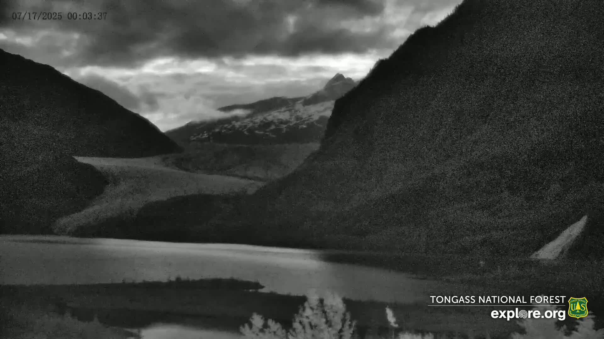 A dark, cloudy night scene shows a calm lake nestled between mountains, one of which has a snow-capped peak.