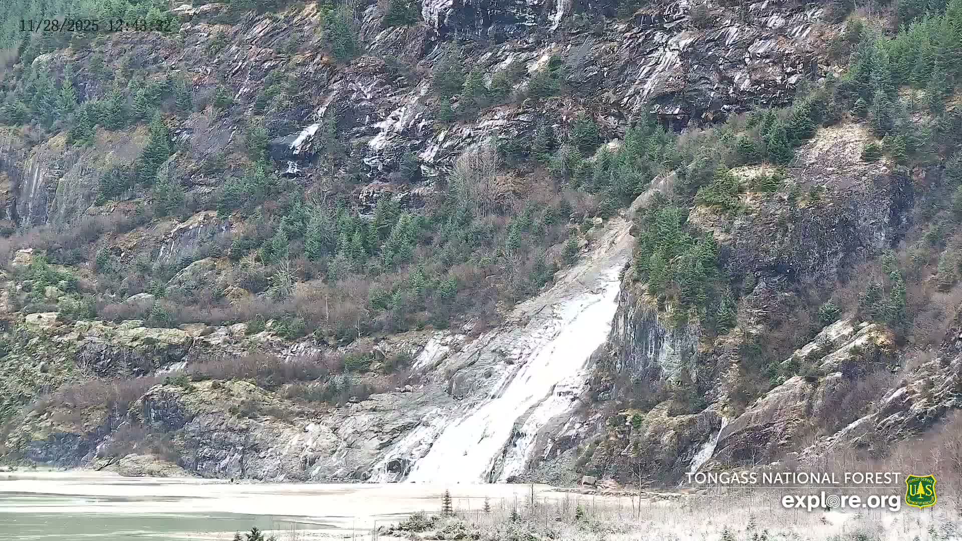 A large waterfall cascades down a steep, rocky mountainside dotted with evergreen trees into a body of icy water, all under clear and cold conditions.