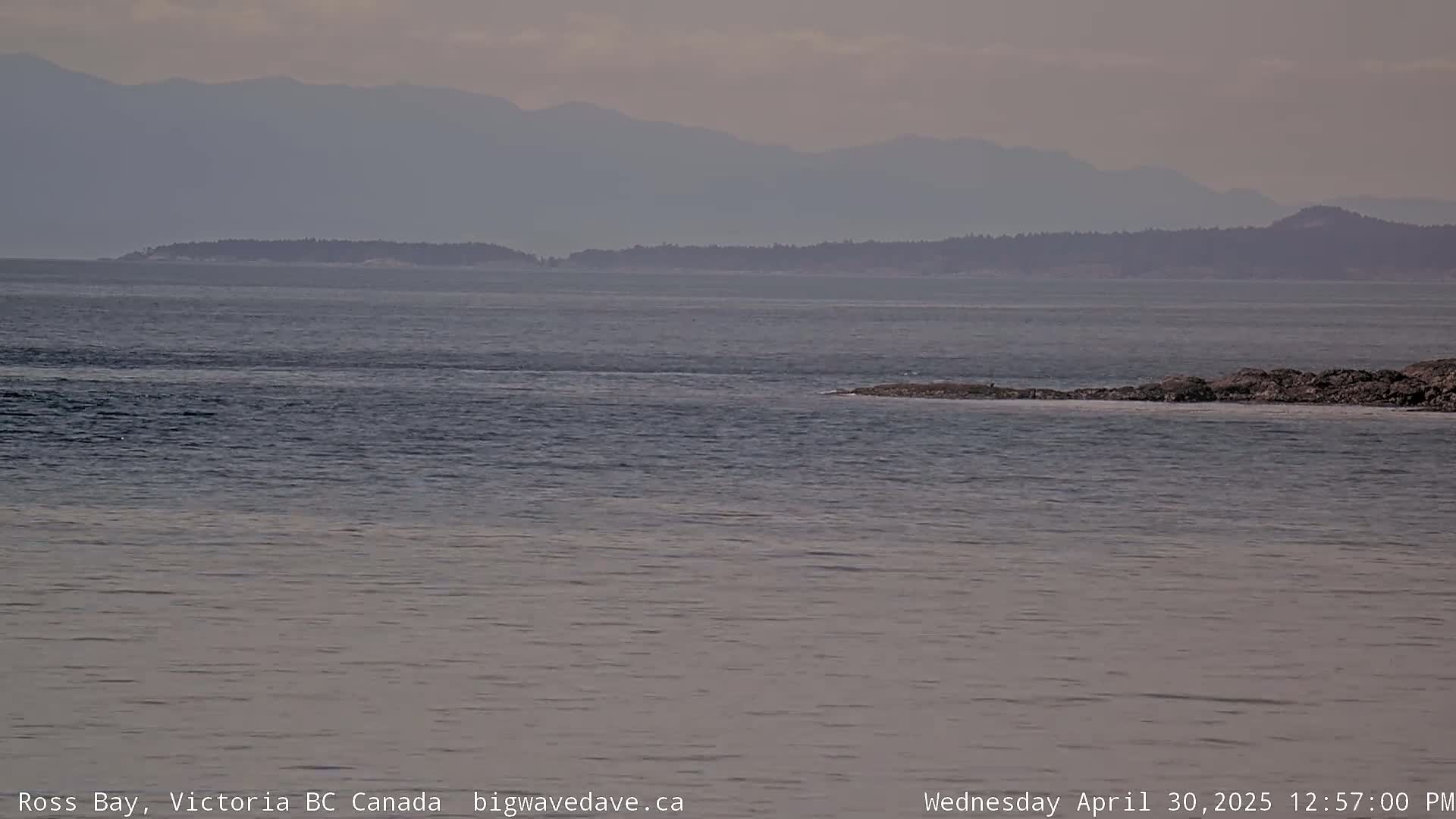 A calm, overcast day reveals a view of a body of water with a distant shoreline featuring low-lying islands and hazy mountains in the background.