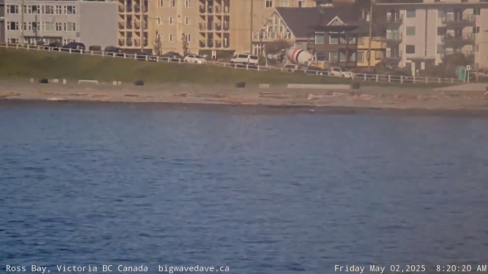 A cement mixer is parked near a beach with calm water and several buildings in the background.