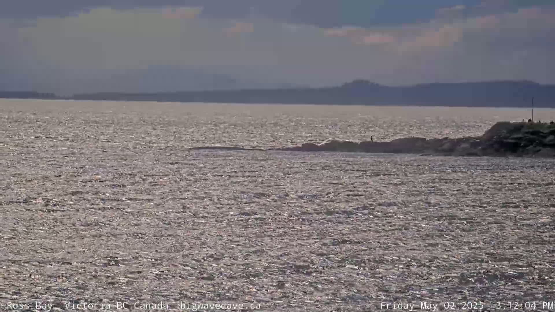 A mostly cloudy day shows a wavy body of water with a rocky shoreline and distant mountains under a partly cloudy sky.