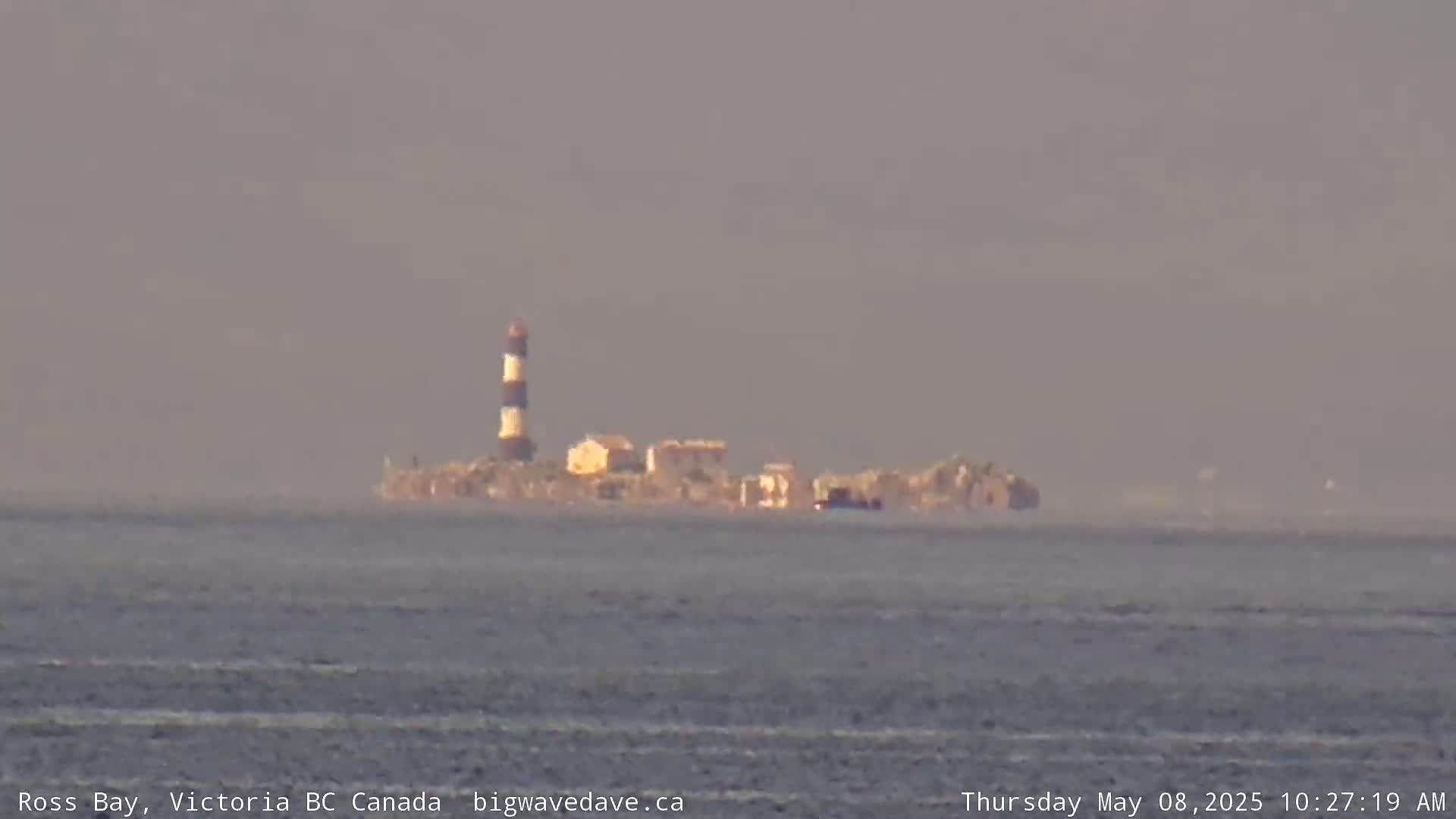A distant island with a lighthouse and several buildings is visible across a calm body of water under hazy conditions.