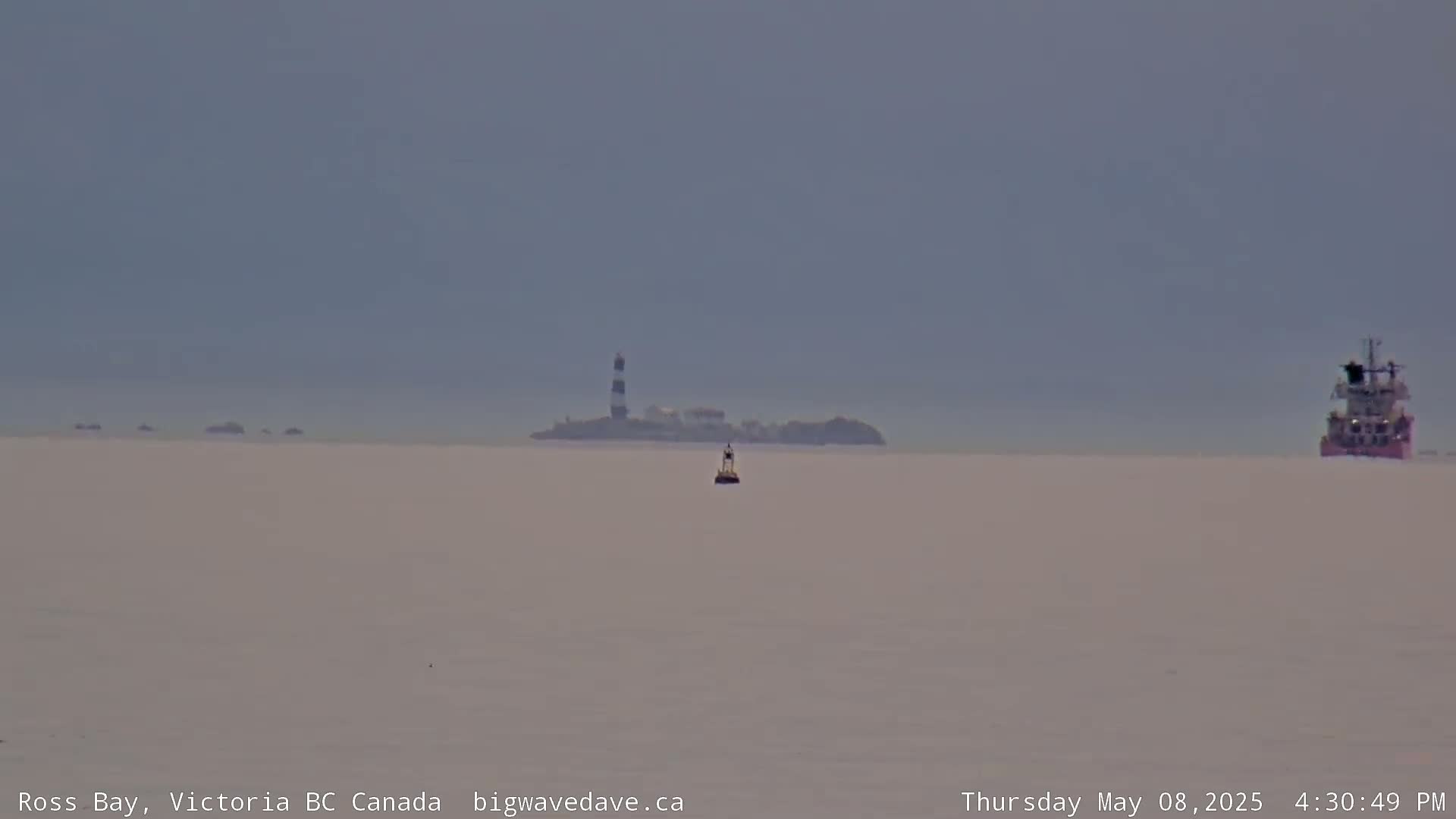 A hazy view of a calm body of water shows a distant island with a lighthouse, a small buoy, and a large ship.