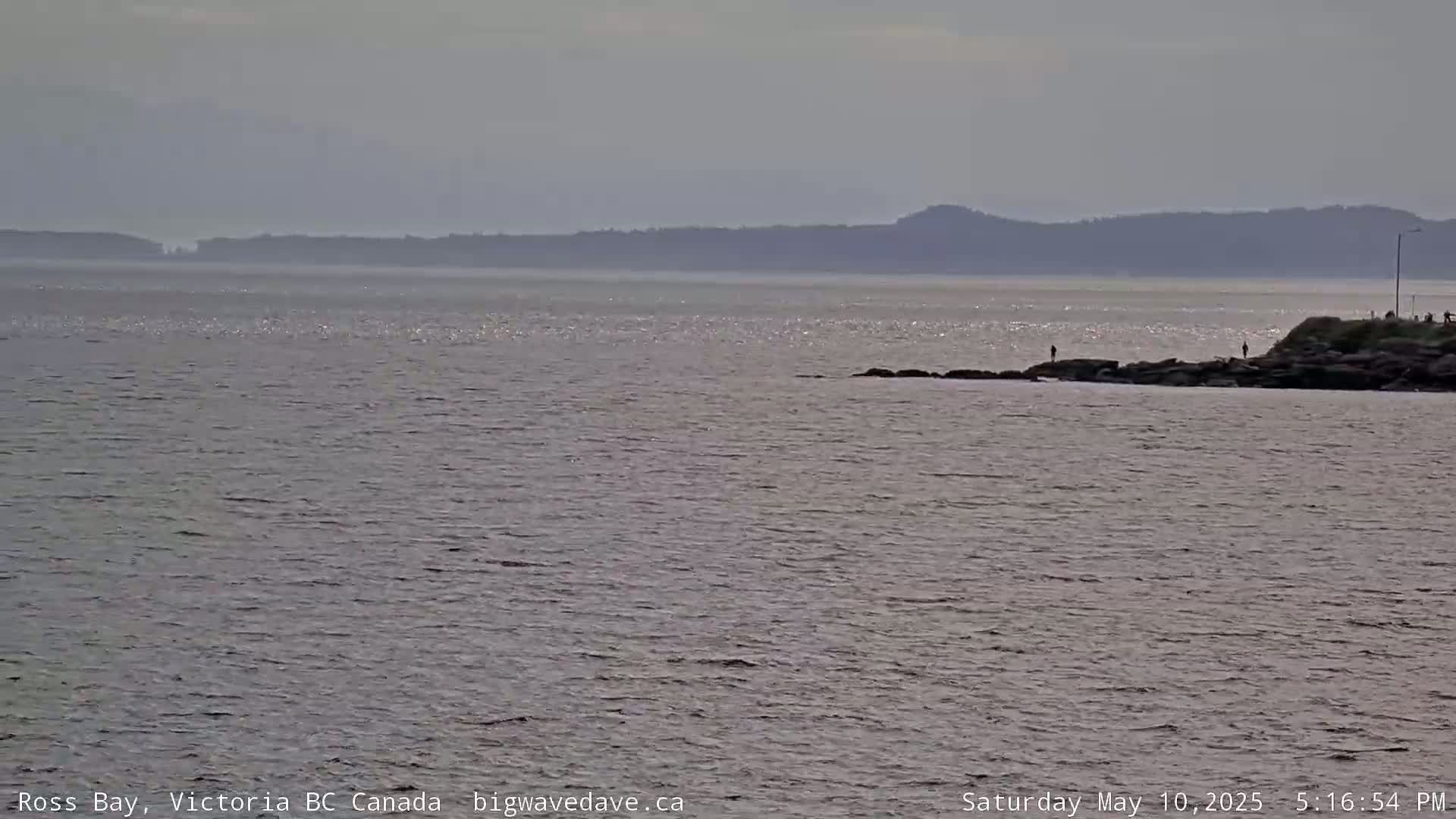 A calm, hazy day shows a body of water with a distant shoreline and a few people on rocks in the foreground.