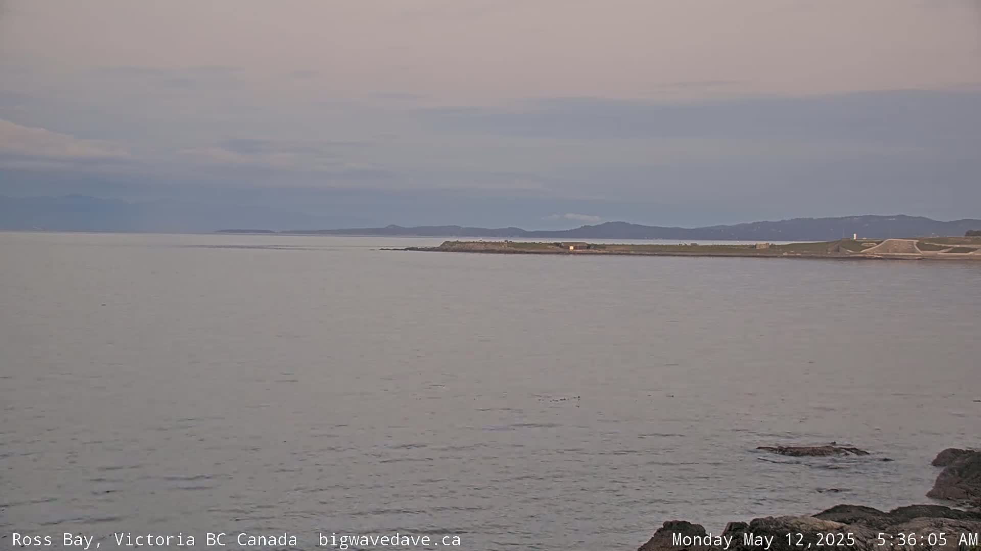 A calm, overcast day shows a wide expanse of grey water with a distant shoreline and mountains under a pale sky.