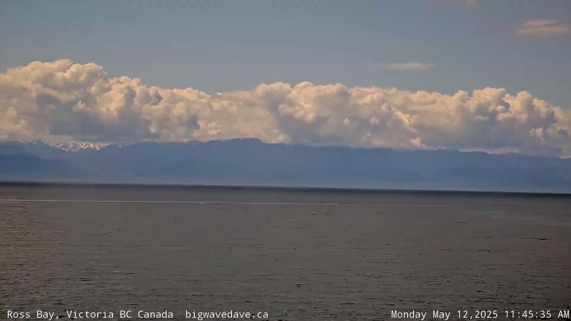 A calm body of water stretches to a distant mountain range partially obscured by a band of fluffy clouds under a mostly clear sky.