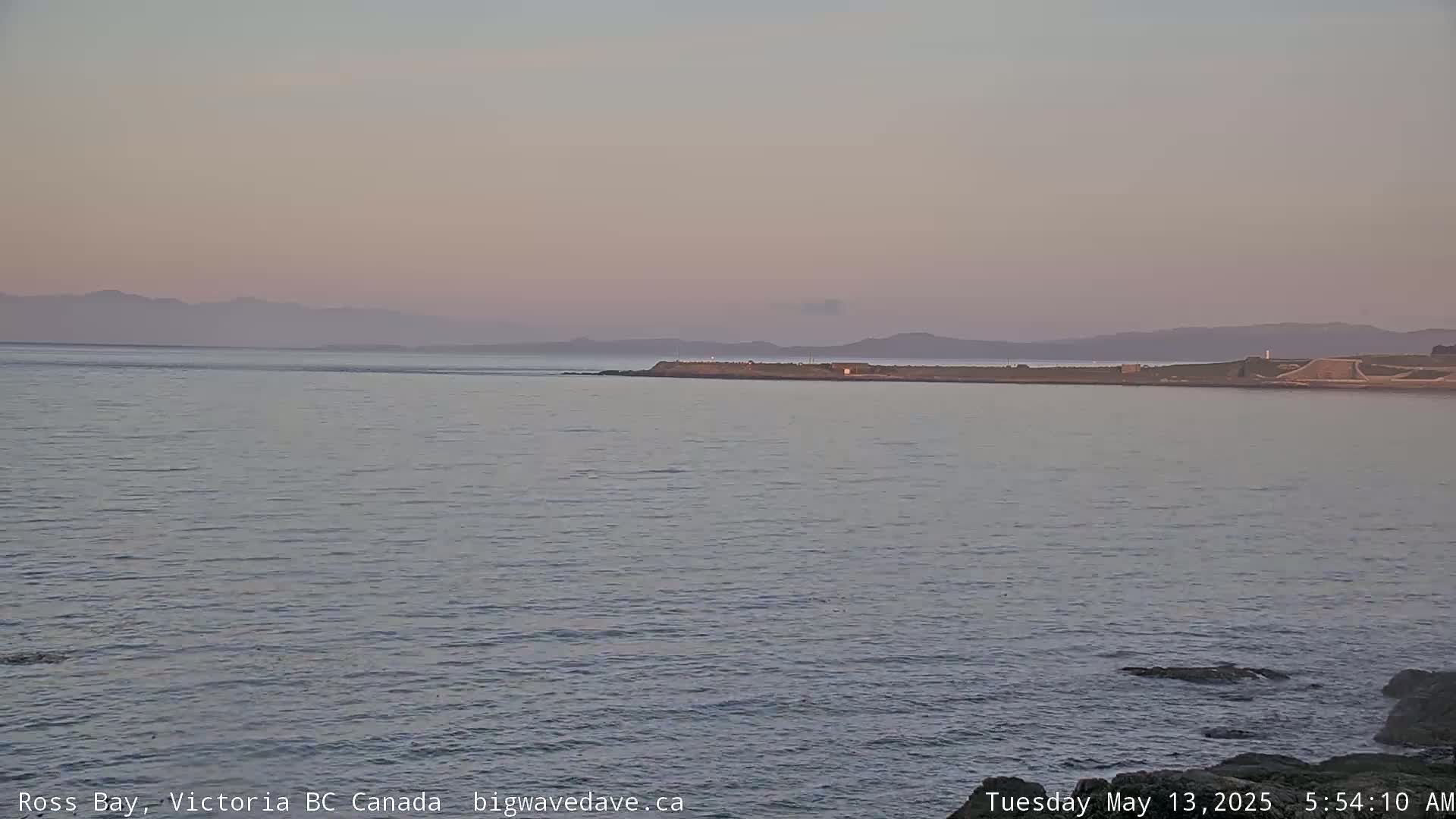 A calm body of water stretches to a distant shoreline under a pale pink sky.