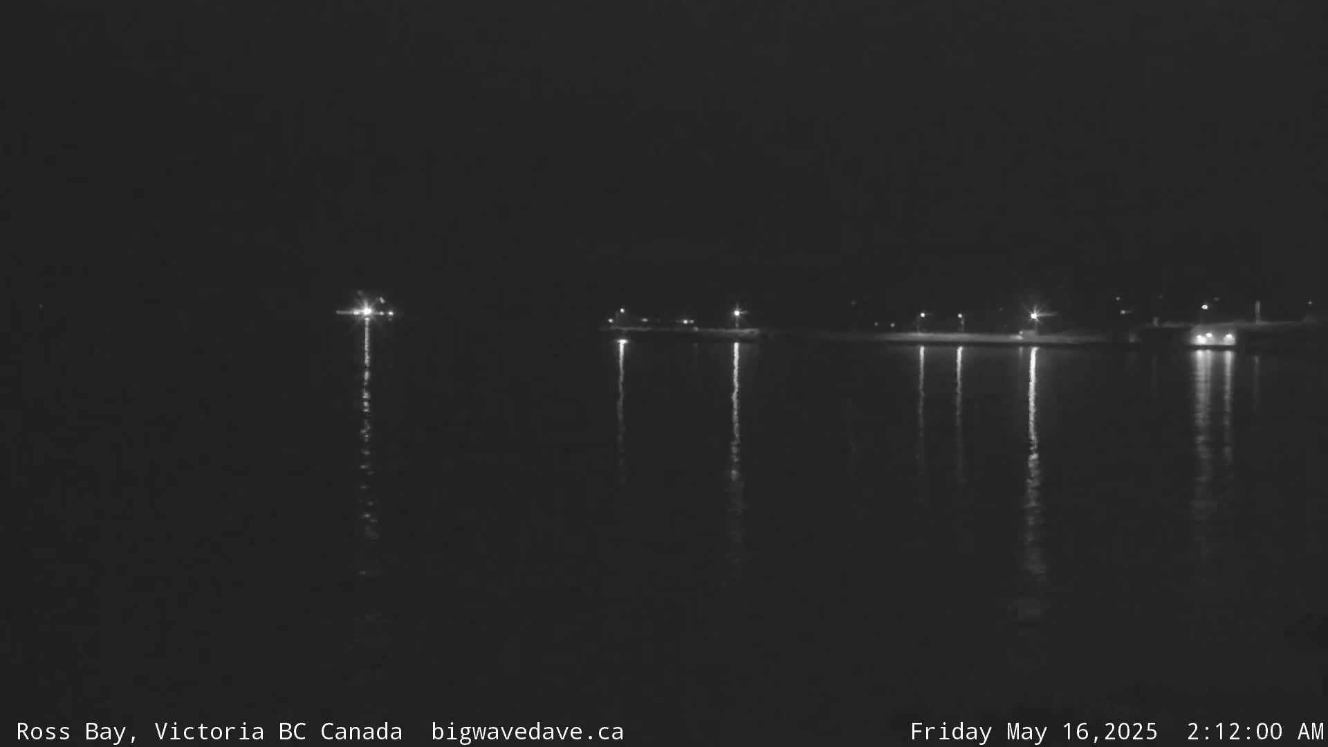 A dark nighttime scene shows a calm body of water reflecting the lights of a distant shoreline.