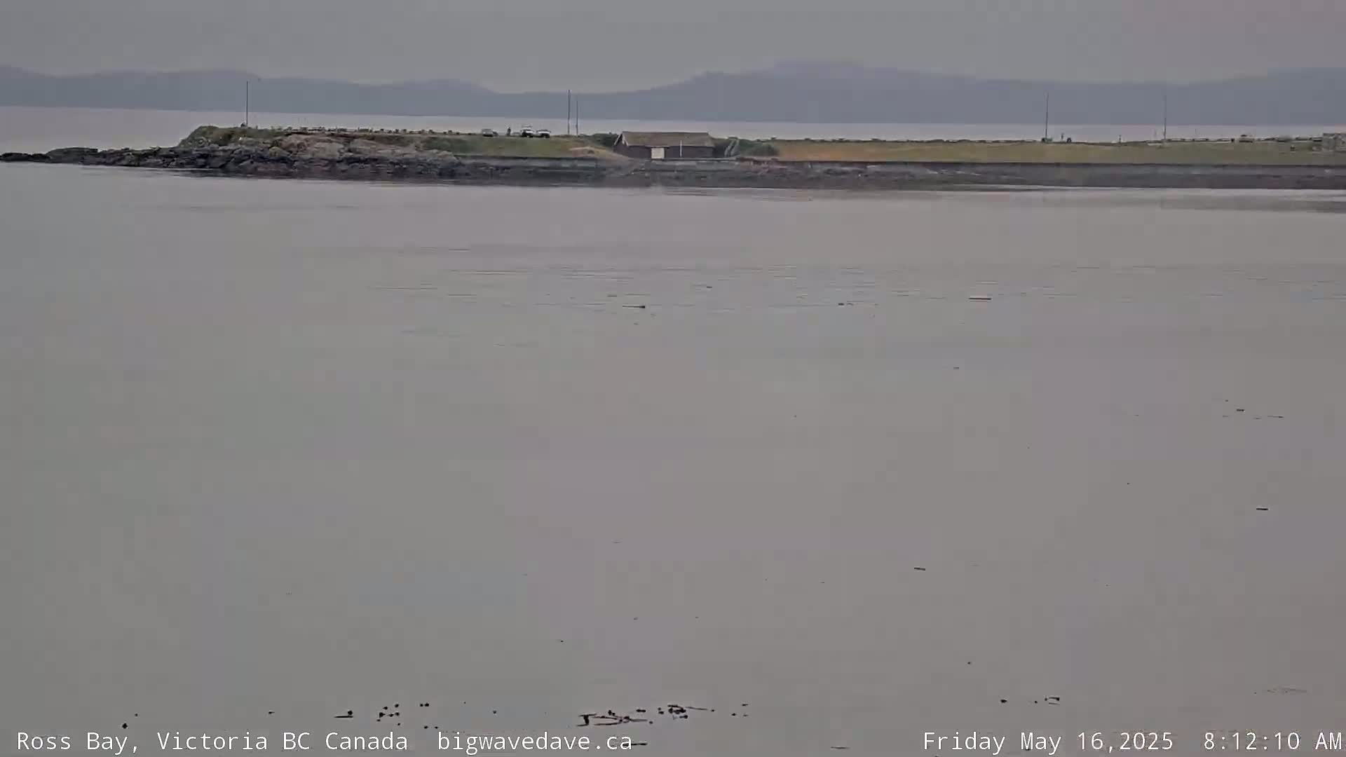 A calm, overcast day shows a rocky shoreline with a small building, a body of water extending to distant mountains.
