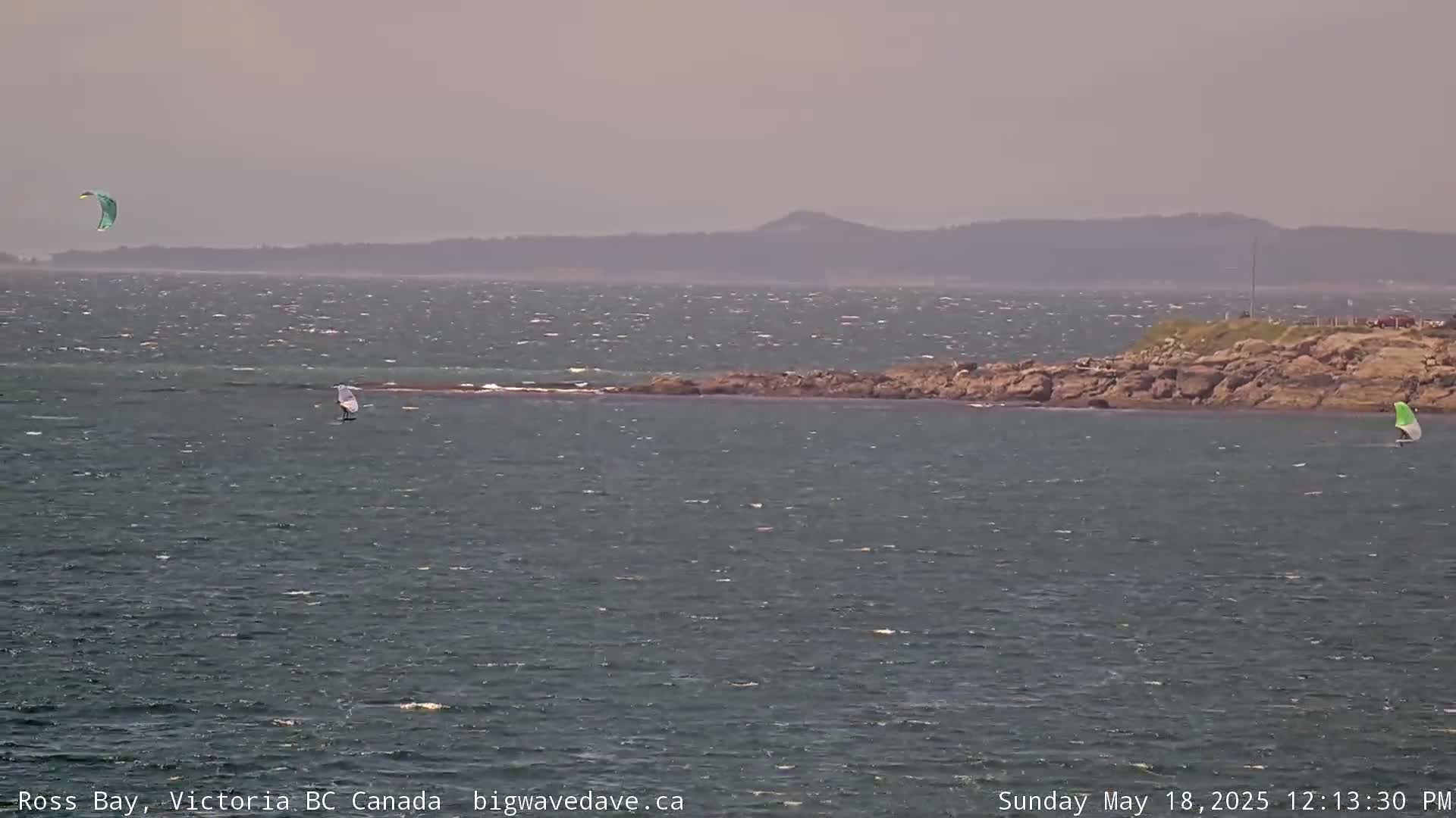 A windy, hazy day shows a kitesurfer and two windsurfers on a body of water near a rocky shore with distant hills.