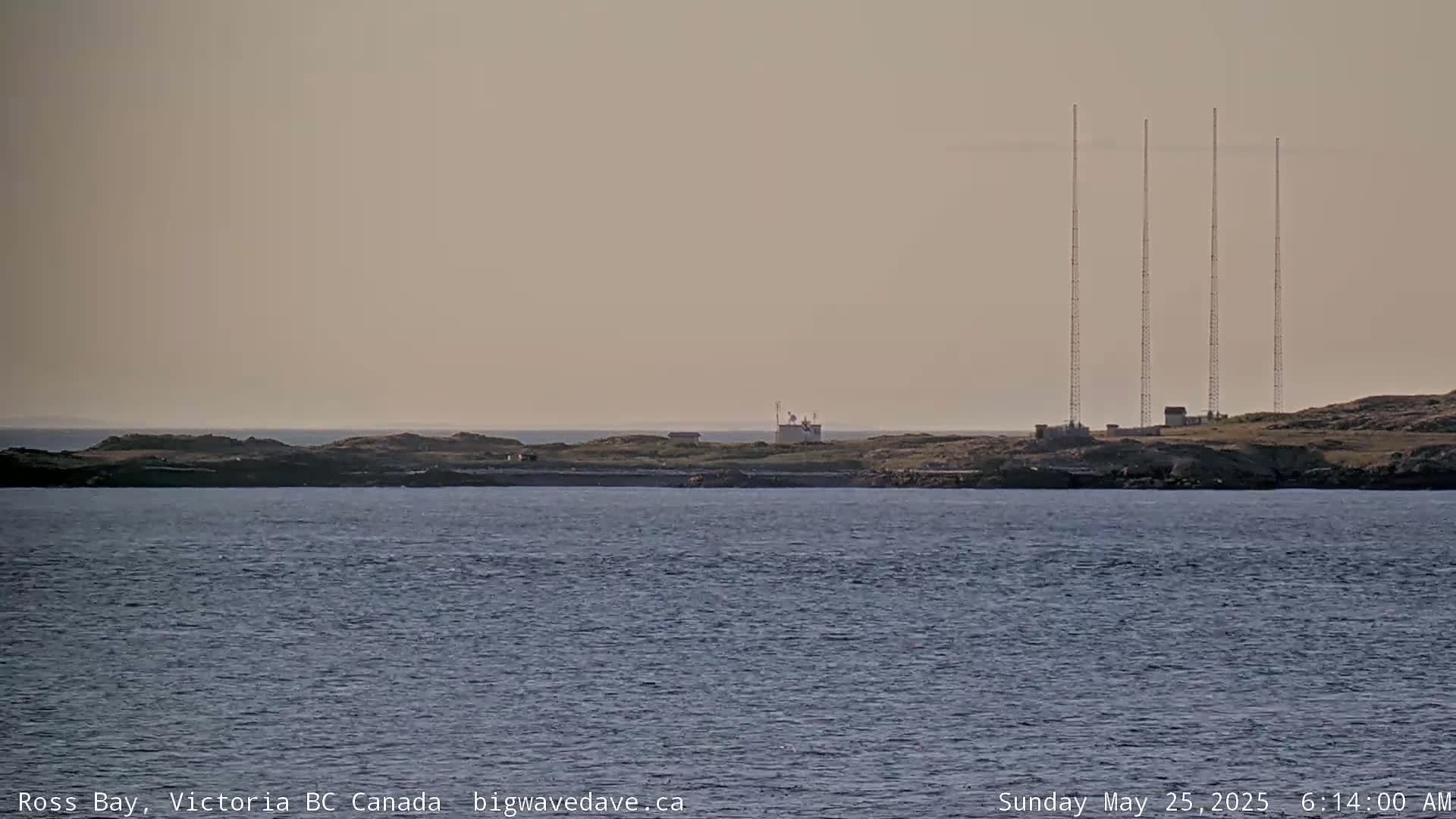 A calm body of water stretches toward a rocky island with several tall antennae and a small building visible under a hazy sky.