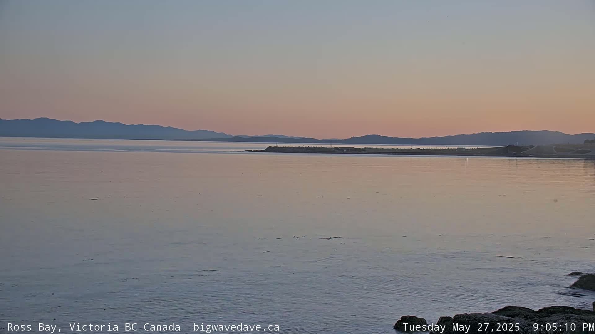 A calm bay at sunset, with a distant mountain range silhouetted against a pale pink sky.
