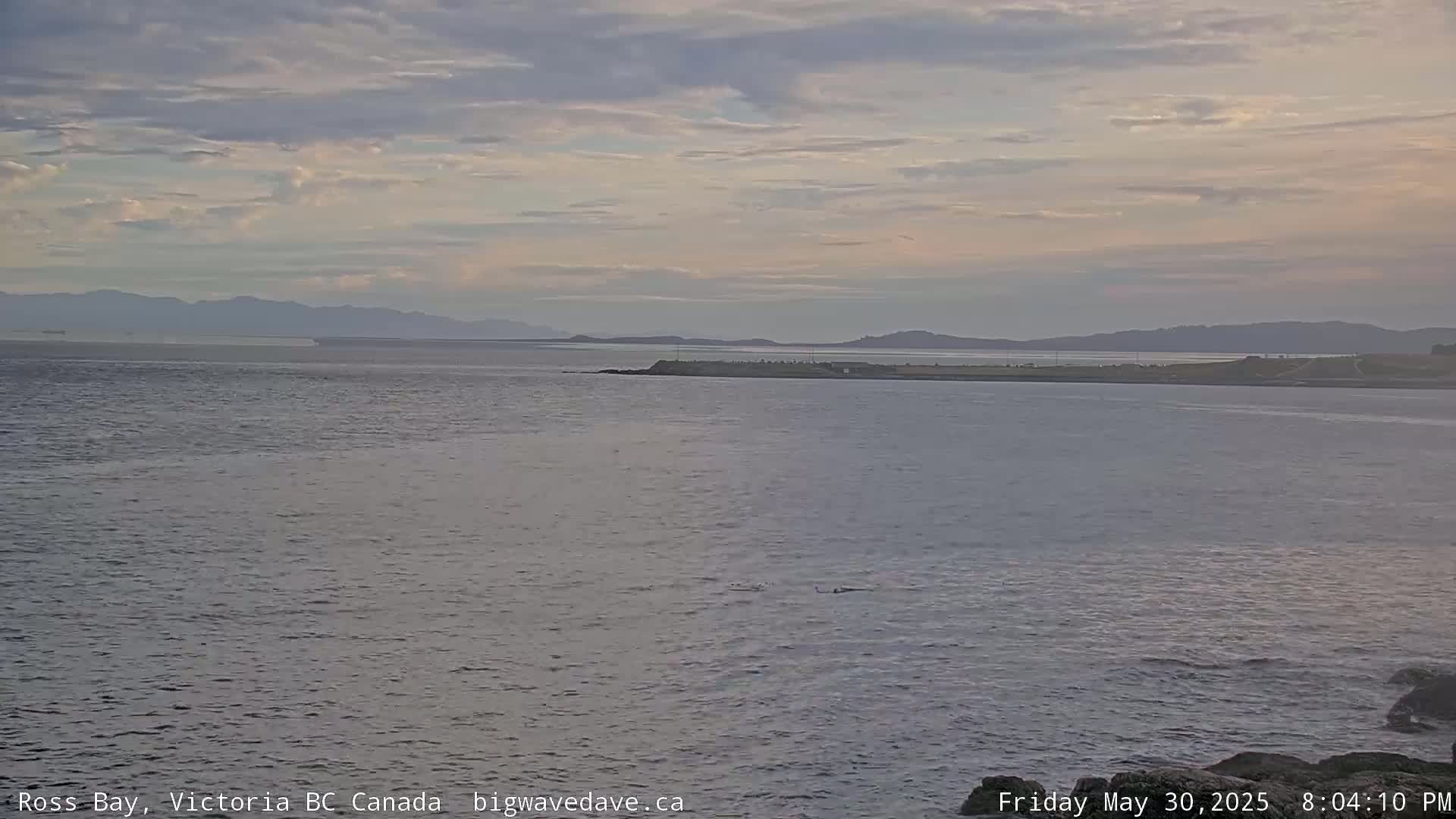 A calm bay with distant mountains under a mostly cloudy, twilight sky.