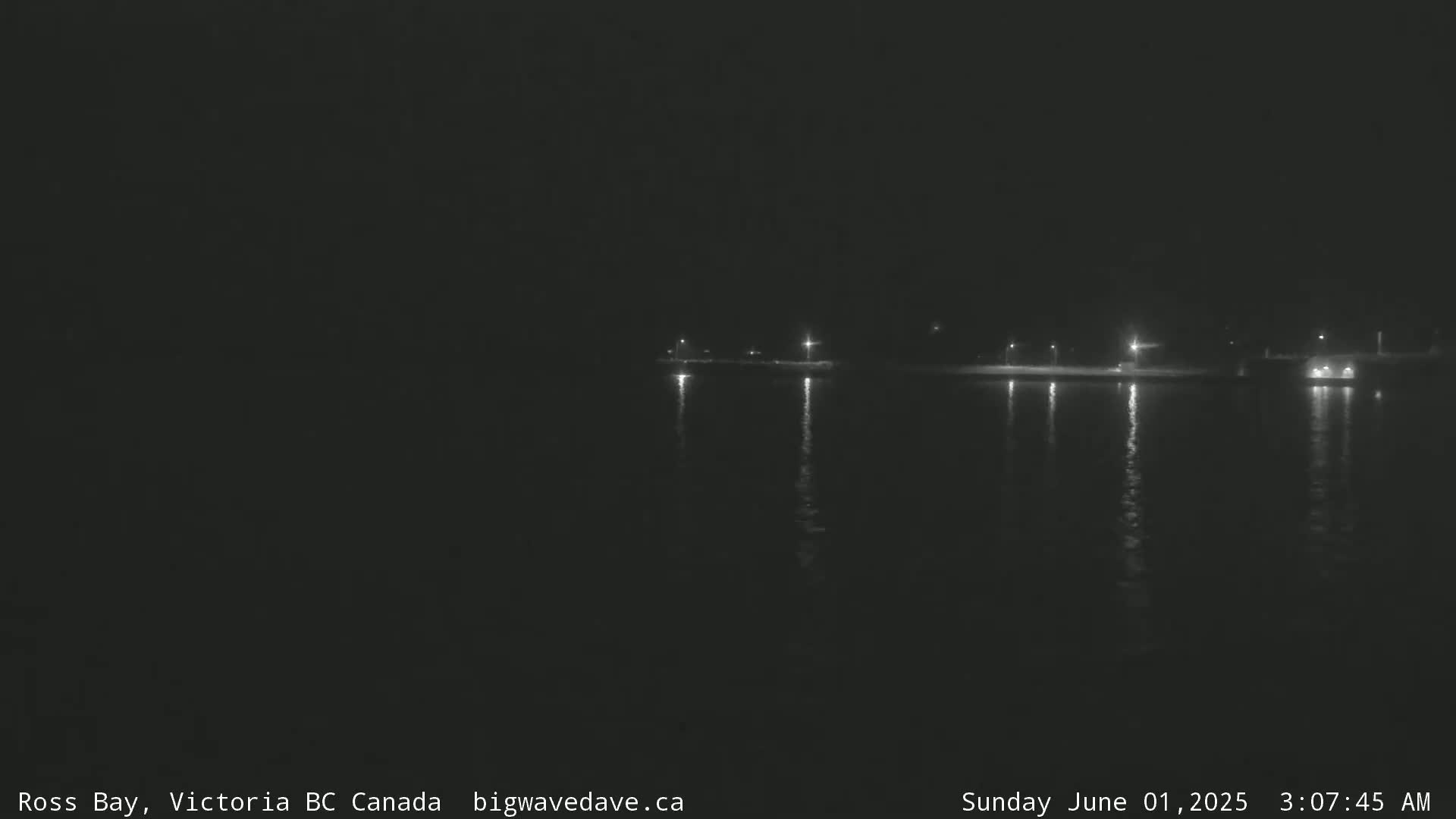 A dark nighttime scene shows a calm body of water reflecting the lights of a distant shoreline.