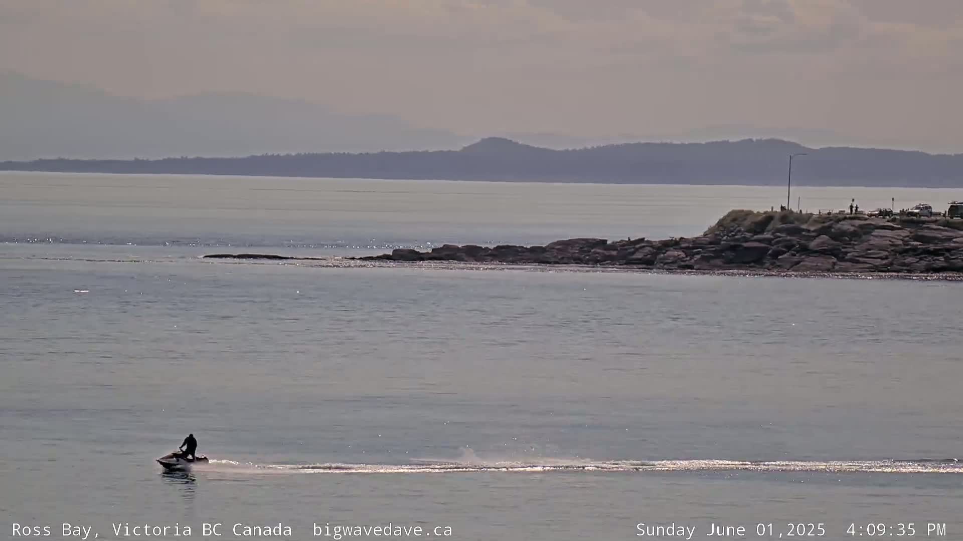 A person on a jet ski is riding across a calm body of water with a rocky shoreline and distant mountains under a mostly clear sky.
