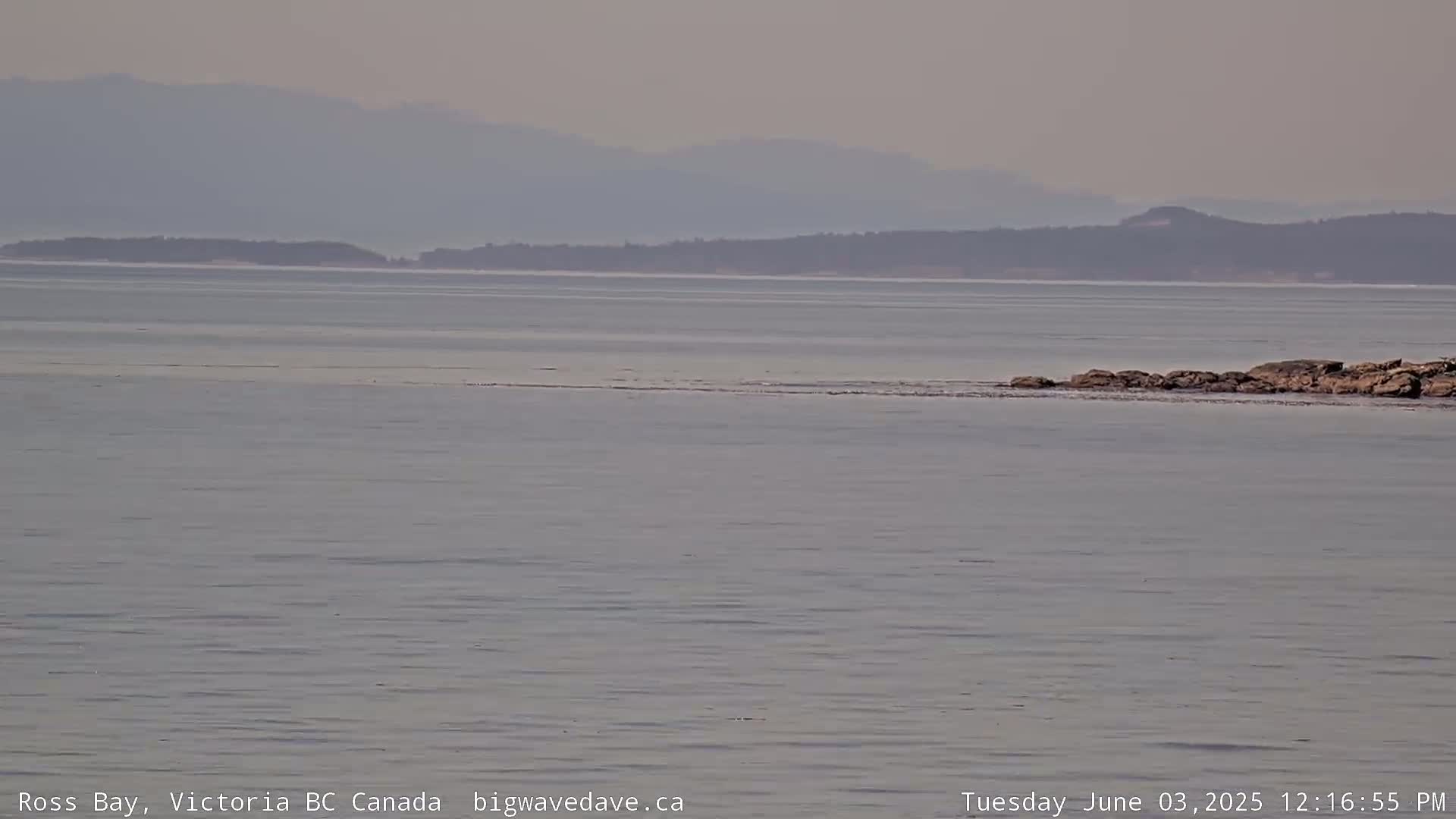 A calm, hazy day shows a view of a body of water with a distant shoreline and mountains under a muted sky.