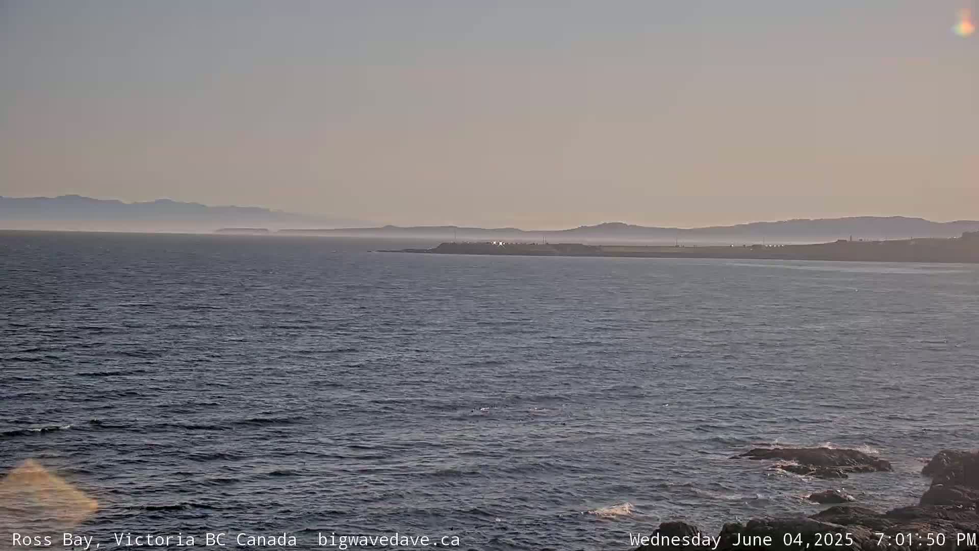 A calm ocean bay stretches to distant hazy mountains under a clear, light sky.