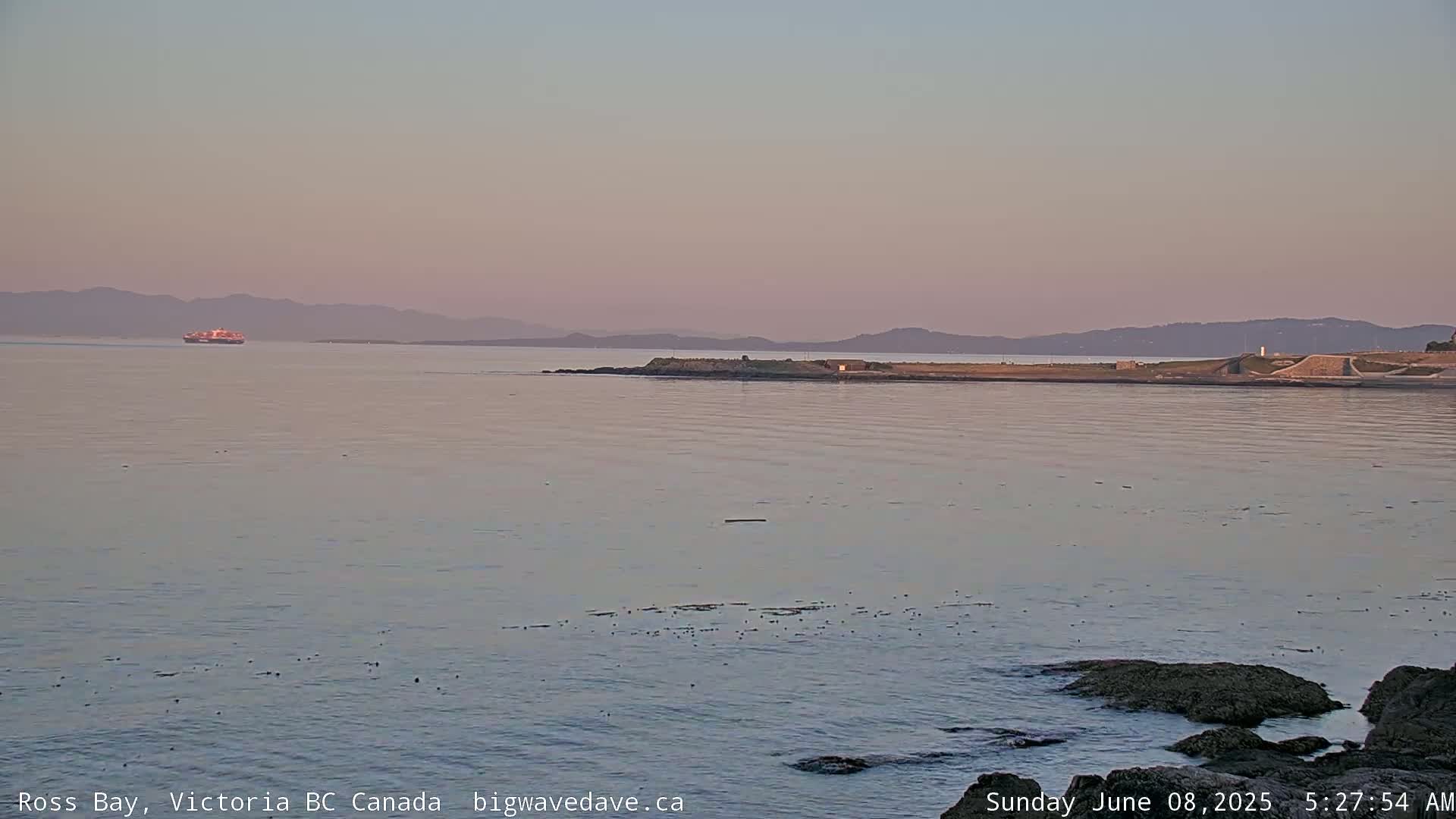 A calm bay at sunset, with a ship in the distance and rocky shoreline in the foreground.