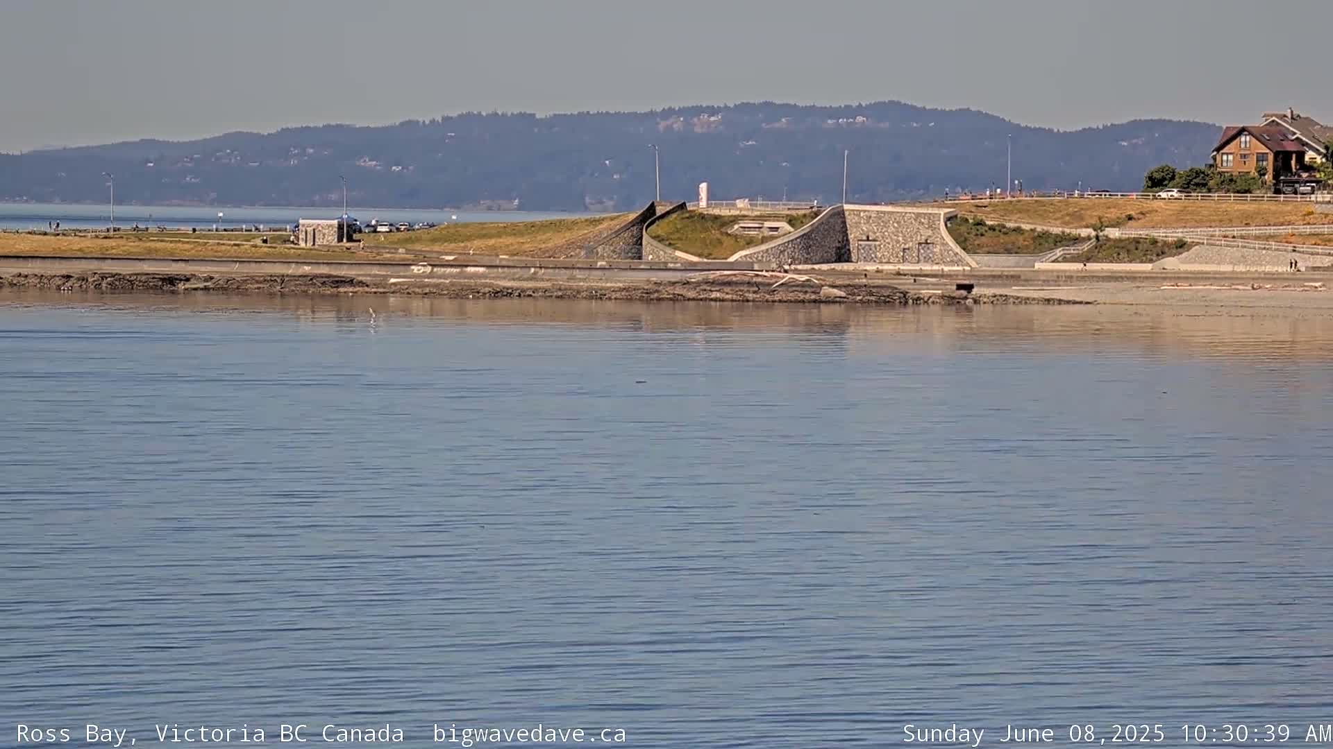A calm body of water stretches out before a shoreline featuring a low stone wall, a grassy area, and a house in the distance under a clear sky.