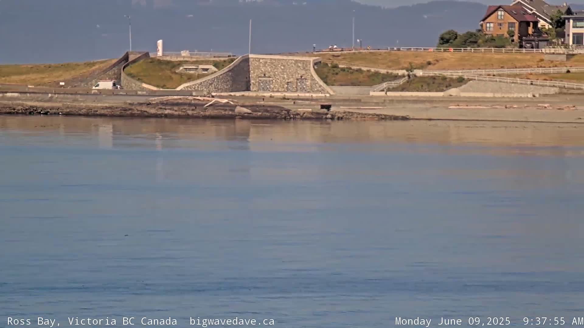 A calm body of water reflects a shoreline featuring a stone wall, a sandy beach, and houses on a hill in the background under a mostly clear sky.