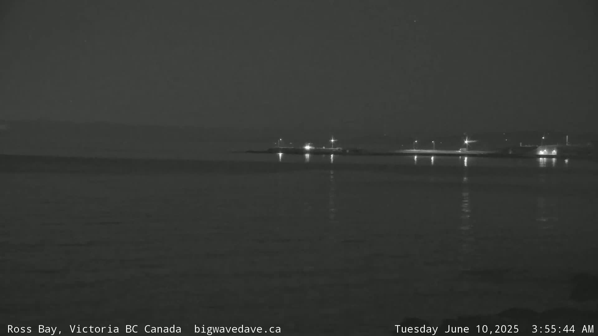 A nighttime, dark, and calm body of water reflects the lights of a distant shoreline.