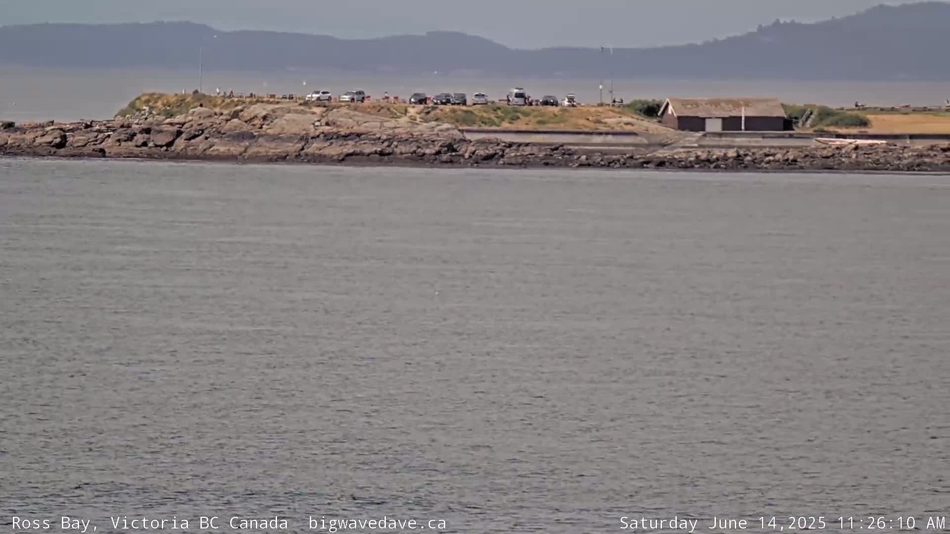 A rocky shoreline with a small building and several parked cars is visible across a calm body of water under a hazy sky.