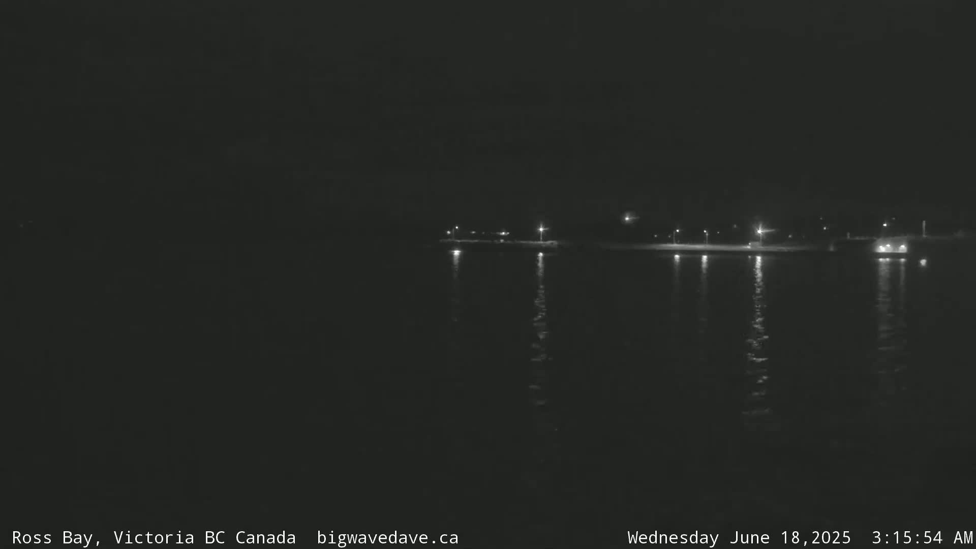 A dark nighttime scene shows a calm body of water reflecting the lights of a distant shoreline.