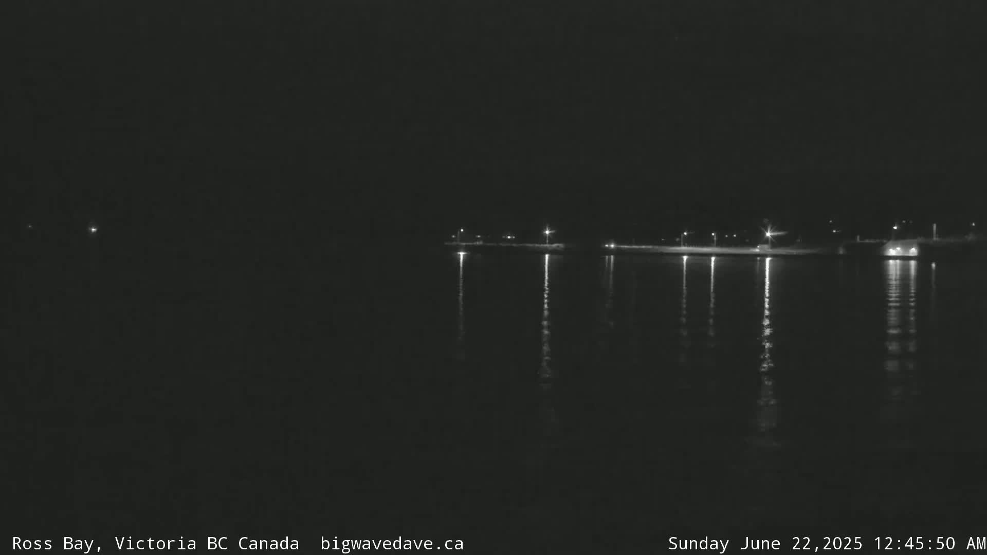 A dark nighttime scene shows a calm body of water reflecting the lights of a distant shoreline.