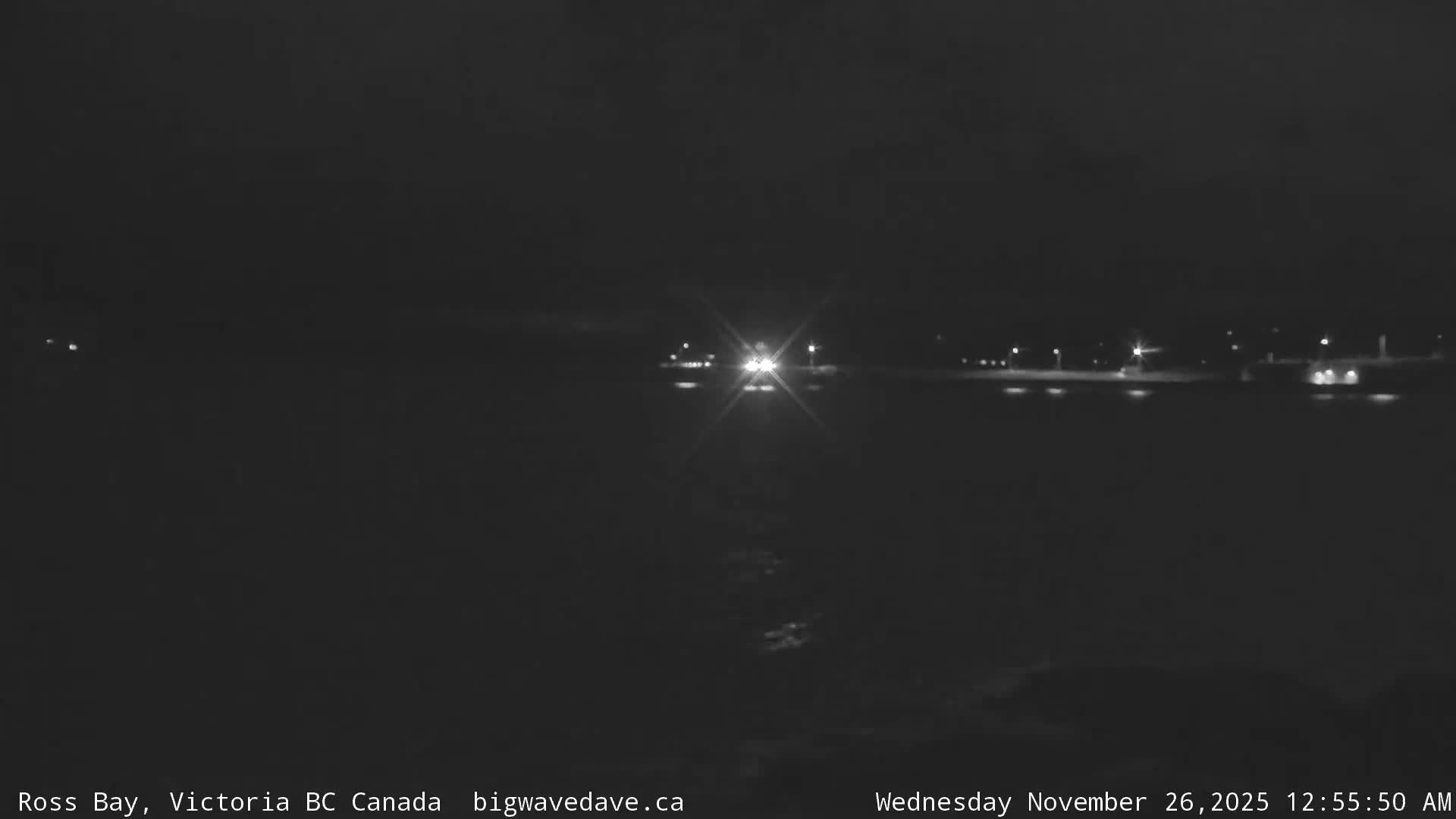 A clear and very dark night scene over calm water features bright headlights from a distant vehicle reflecting on the surface, accompanied by other scattered shoreline lights.