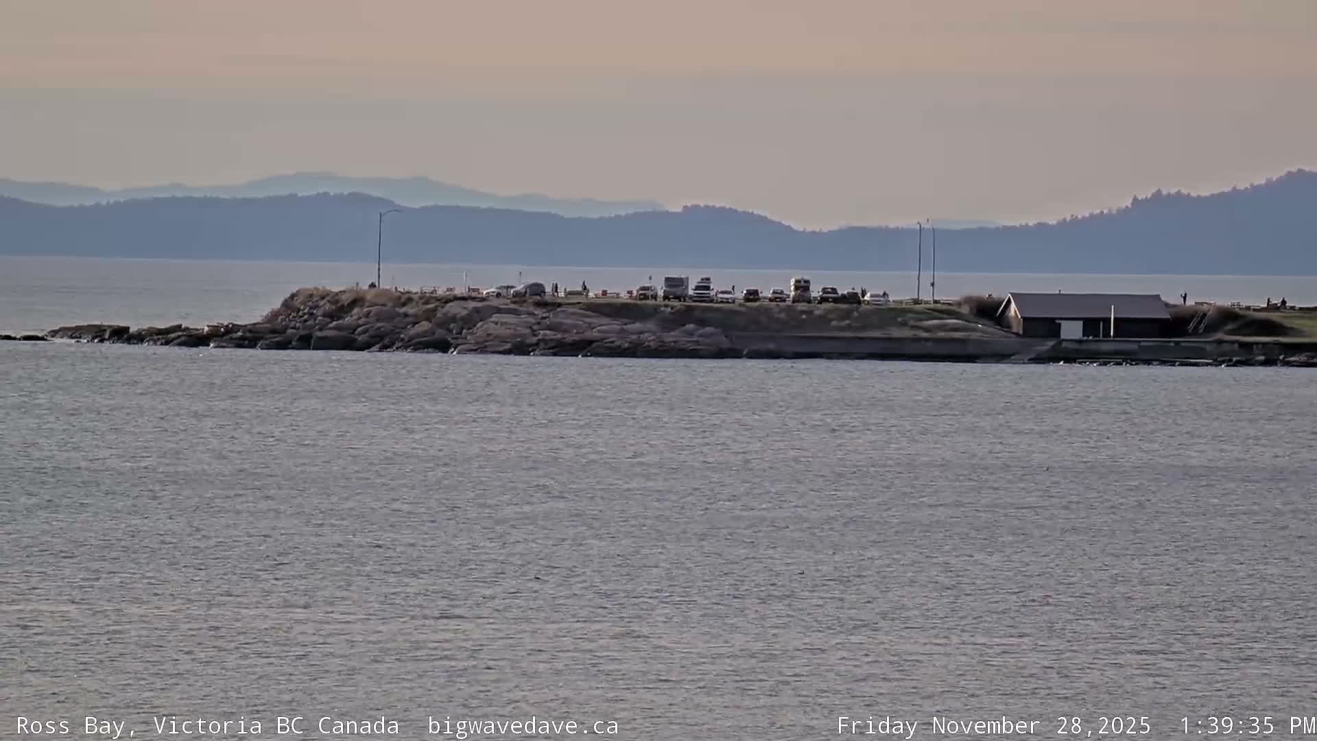 Under an overcast sky, a calm, grey sea laps at a low, rocky peninsula featuring a parking area with vehicles and a small building, with hazy mountains visible in the distant background.