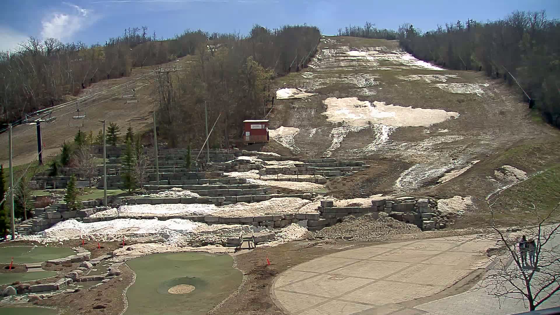 A ski hill with melting snow patches under a sunny sky, showing a chairlift and mini-golf course below.