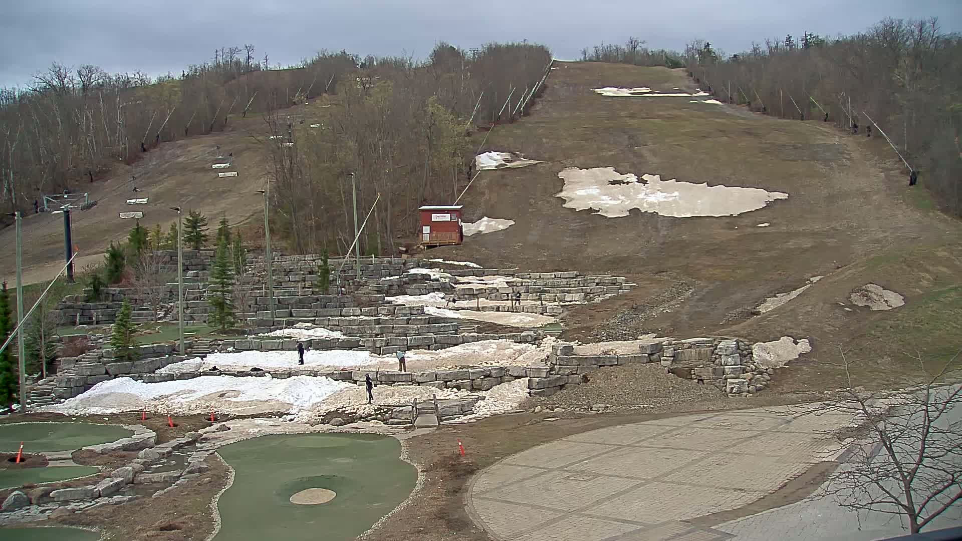 A mostly bare ski slope with remnants of snow, a mini golf course below, and a paved area at the bottom, all under an overcast sky.