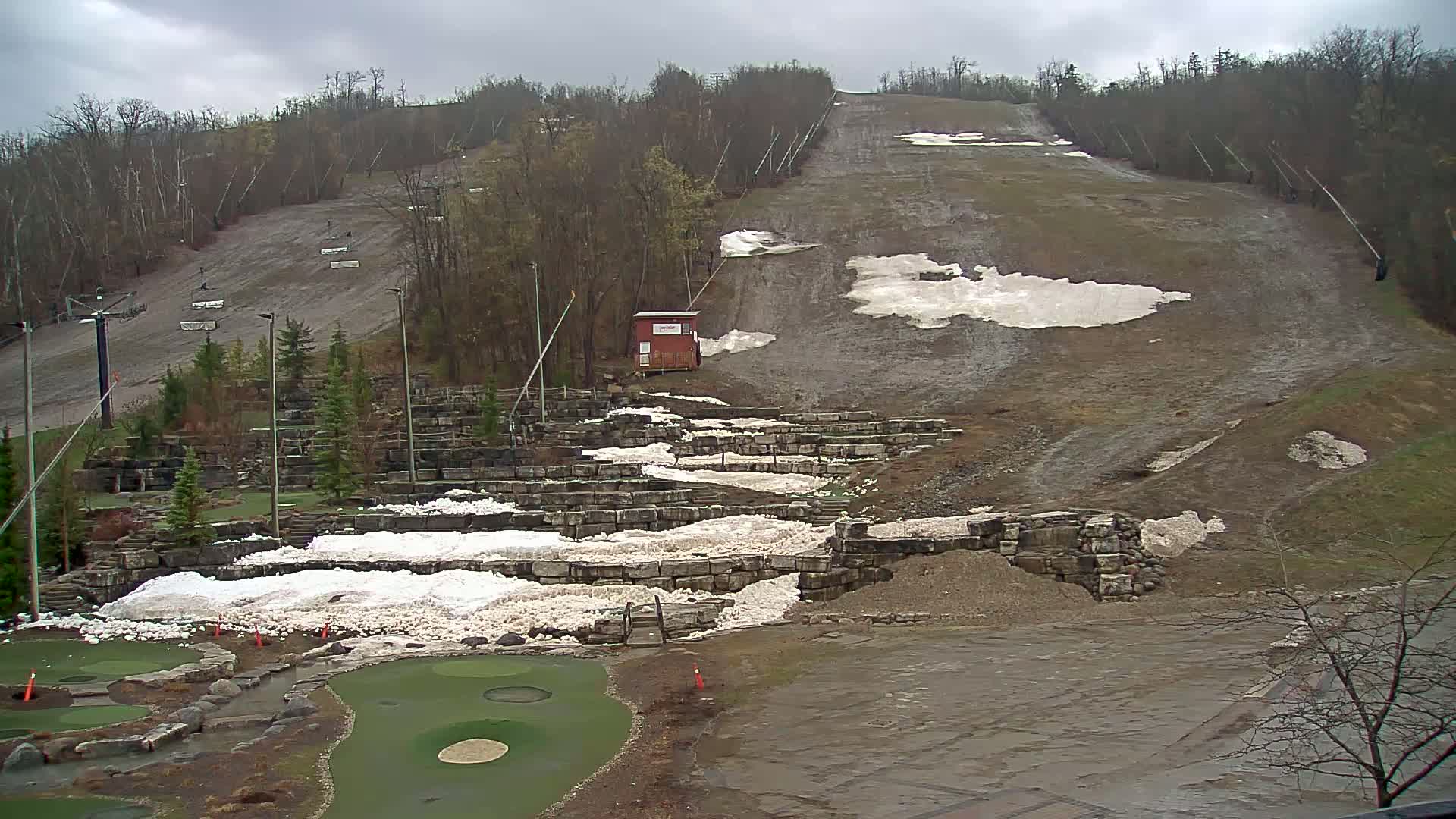 A ski hill with patches of melting snow under an overcast sky, alongside a mini-golf course also with snow remnants.