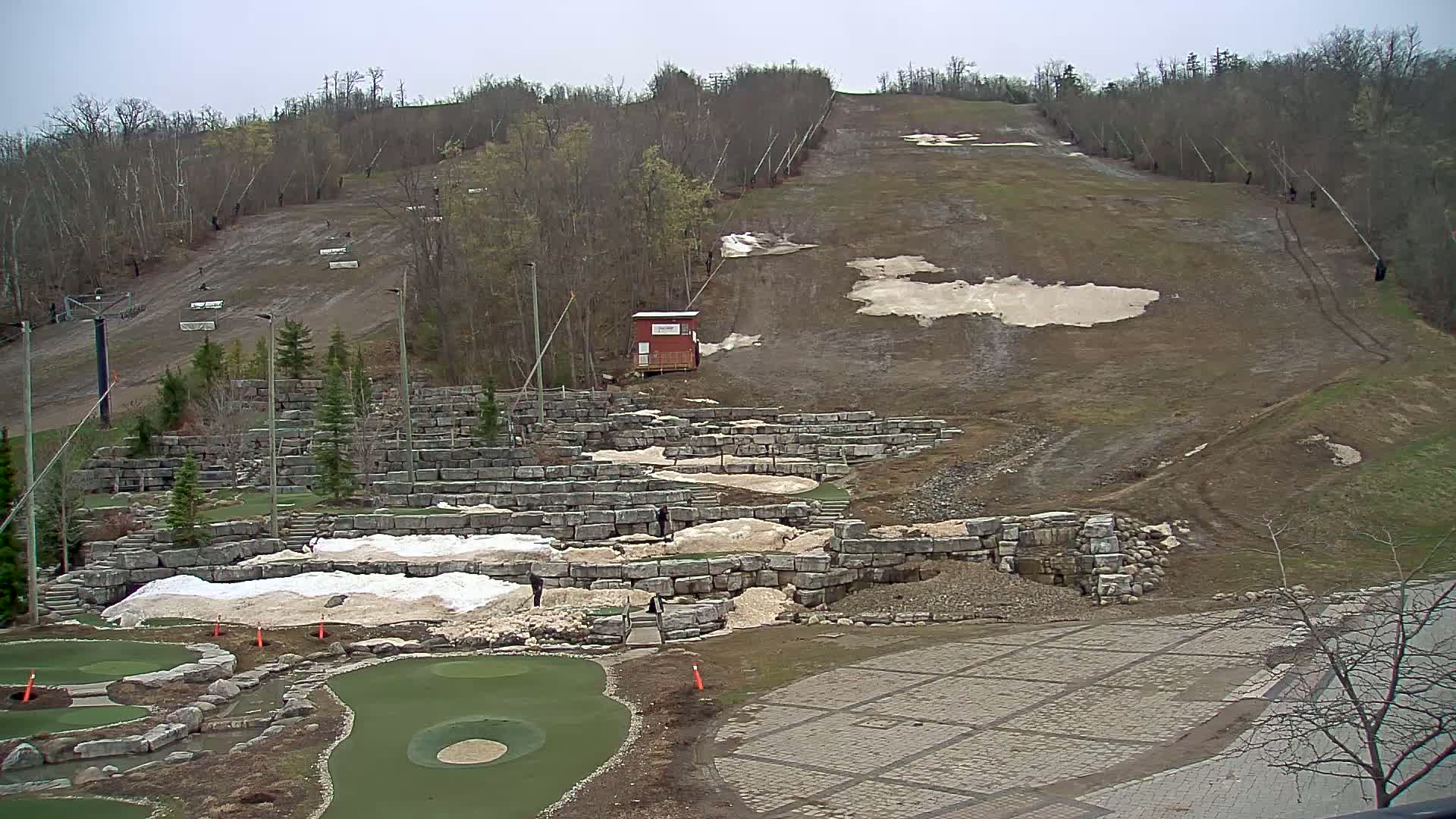 A mostly barren ski slope with remnants of snow and a mini golf course below on a cloudy day.
